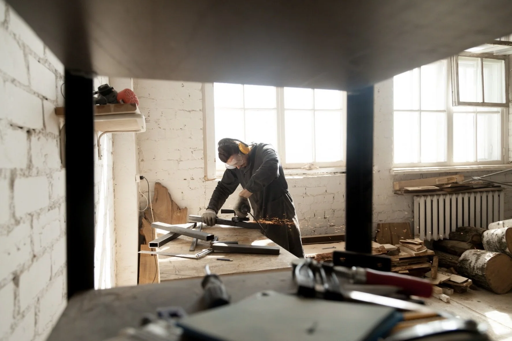 Person using a power tool to cut metal in a bright workshop with large windows, white brick walls, and wood logs.