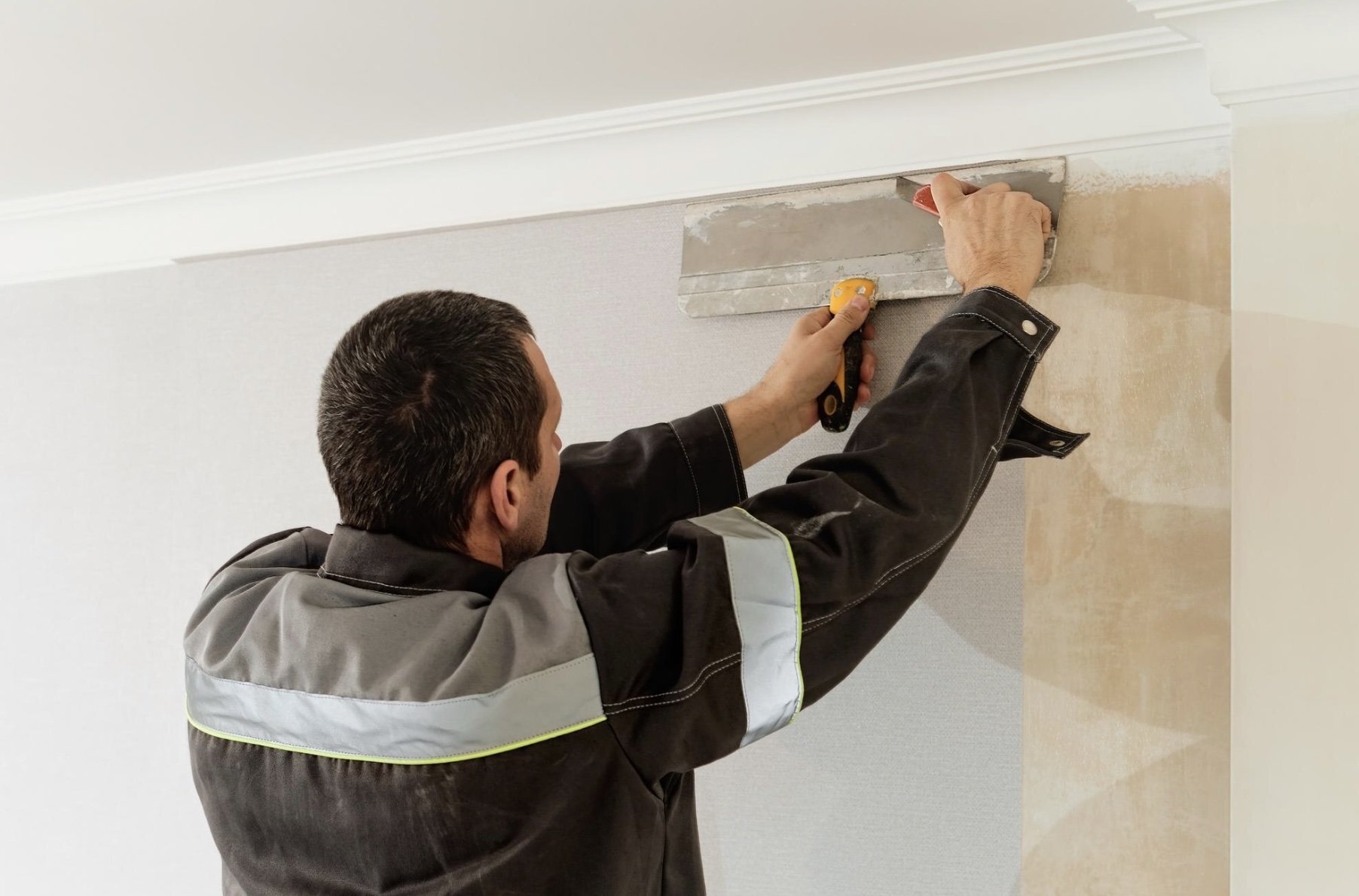 A man installing or repairing a crown molding on the ceiling, holding a utility knife and a trowel, on a white wall.