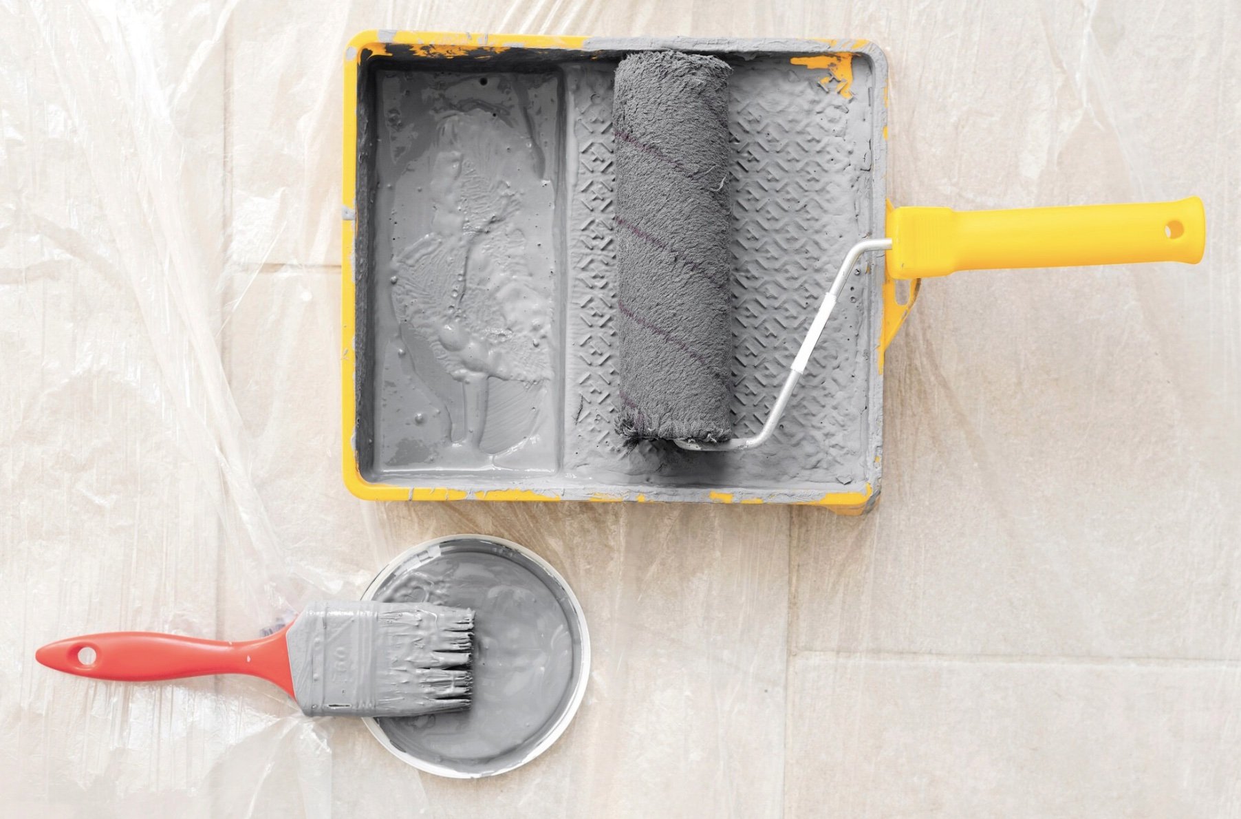A yellow paint roller tray with gray paint, a paint roller with a gray handle and gray cover resting in the tray, a paintbrush with a red handle and gray bristles, and a small container of gray paint on a tiled floor.