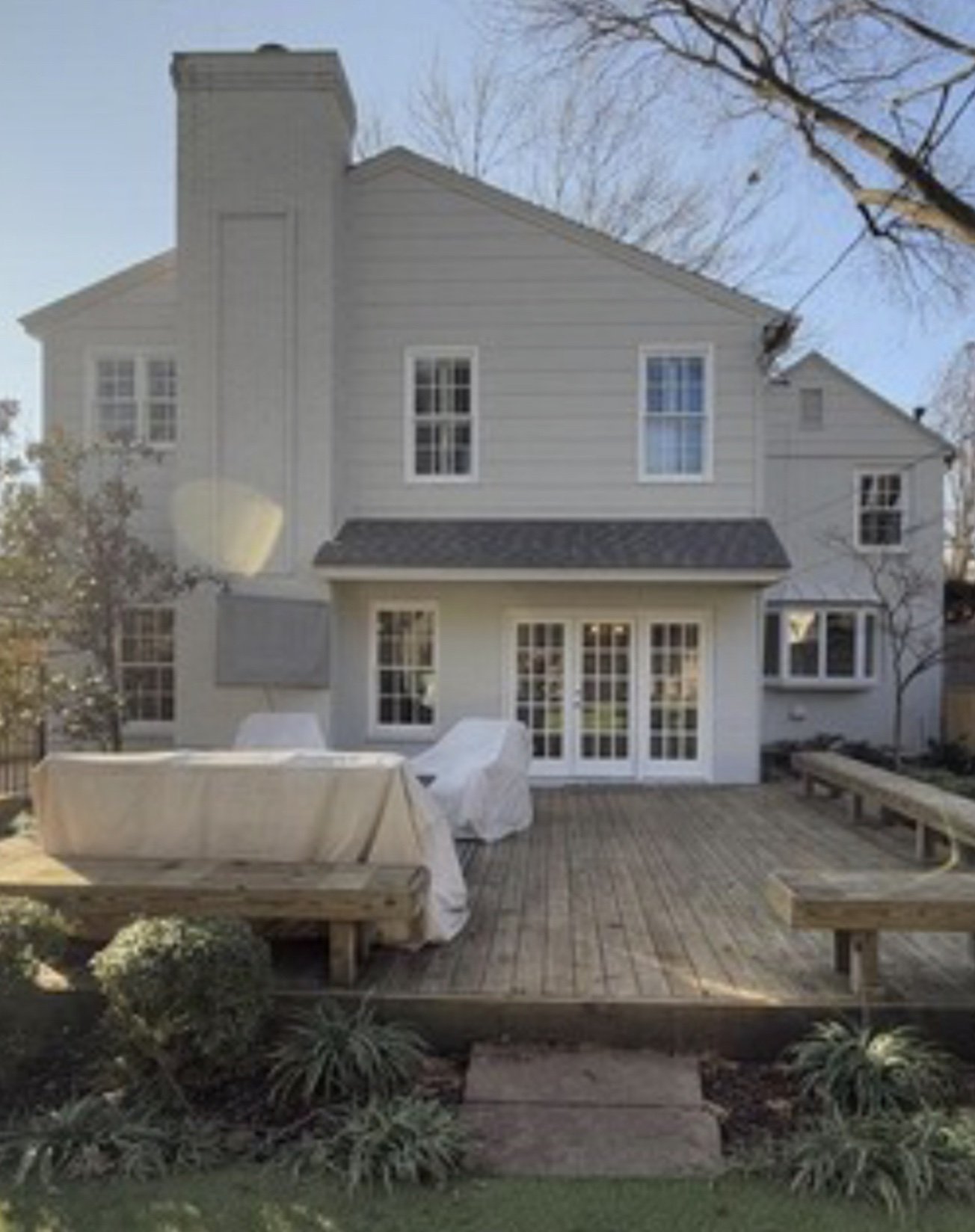 Back view of a two-story white house with a wooden deck, covered patio furniture, and a few small bushes in the garden, with trees and a clear sky in the background.
