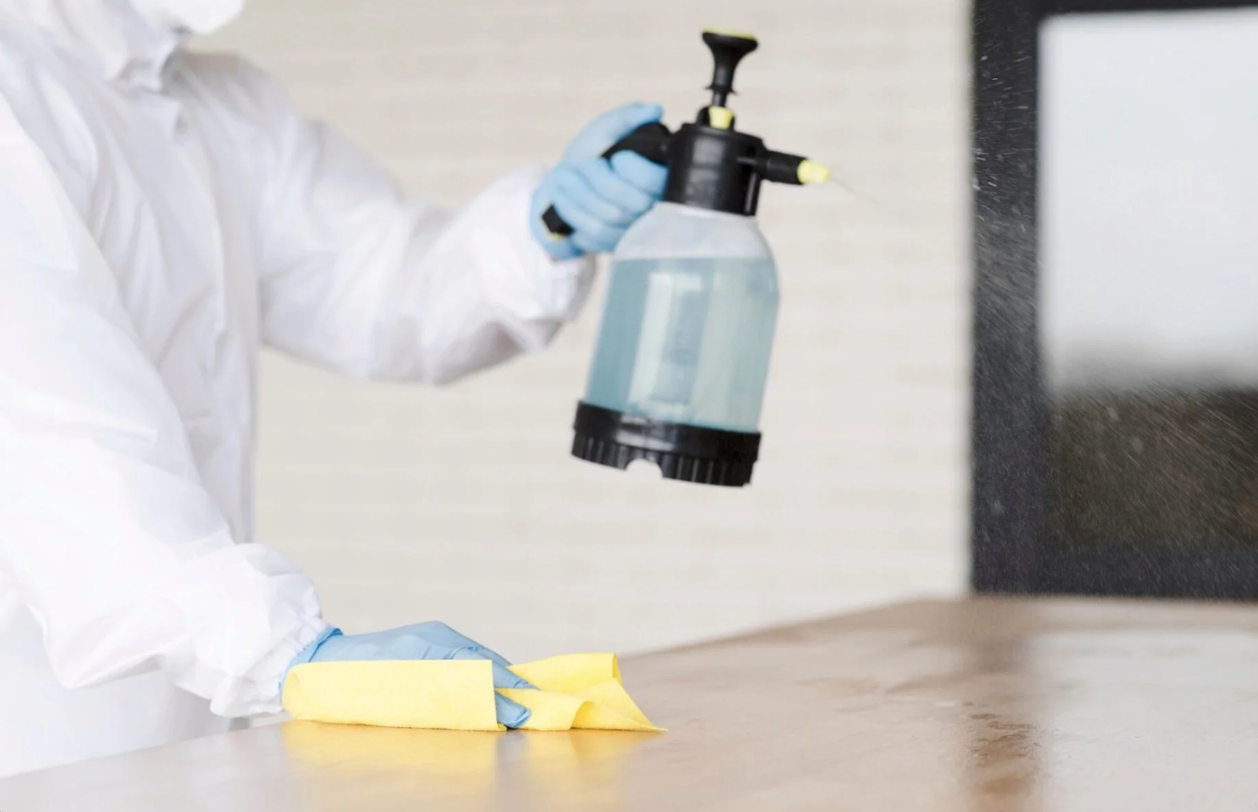 Person wearing white protective suit and blue gloves disinfecting a wooden surface with a spray bottle.