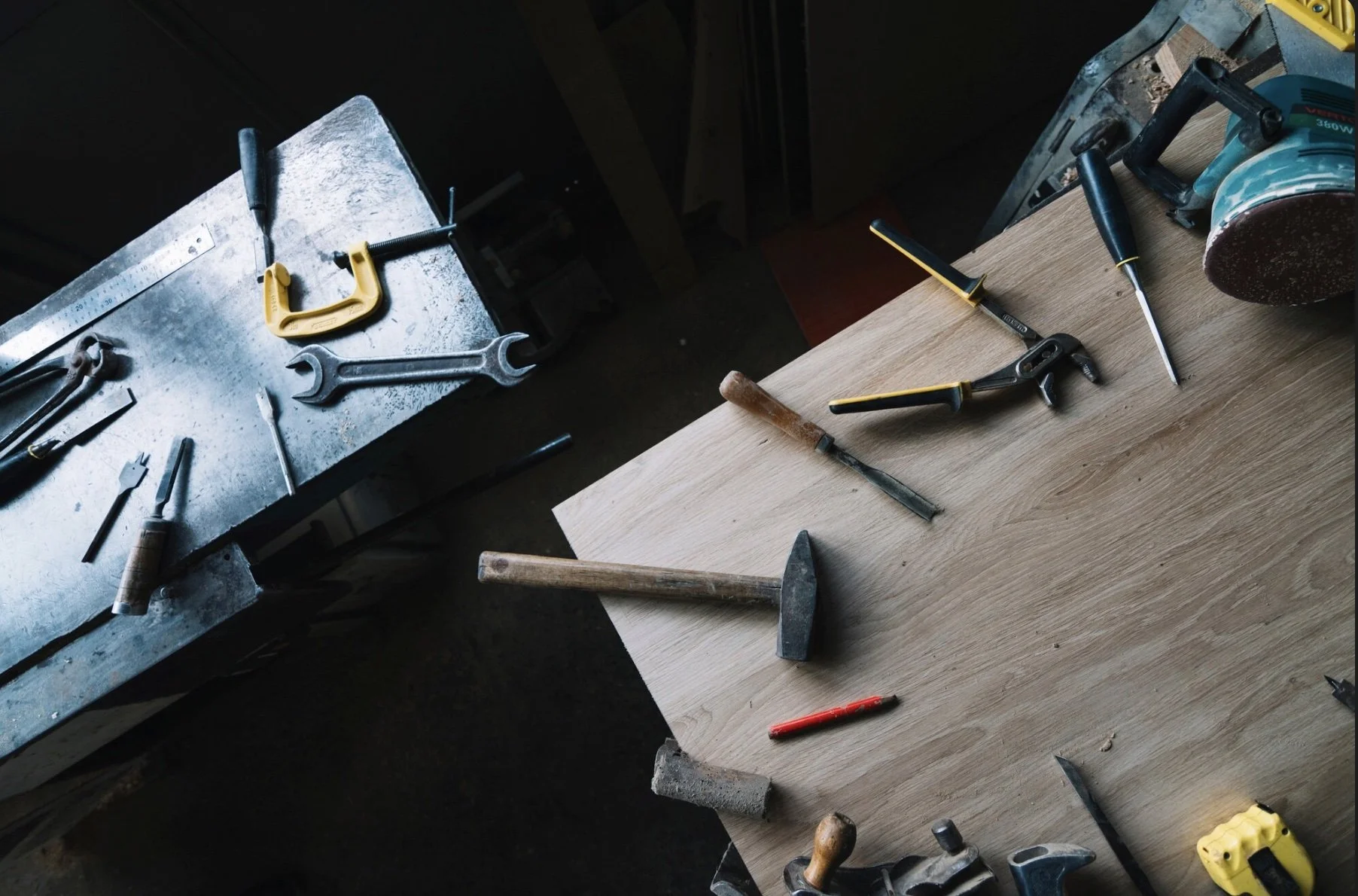 A woodworking workshop with various hand tools including hammers, screwdrivers, chisels, a handsaw, clamps, and a level scattered on a wooden workbench and a metal table.