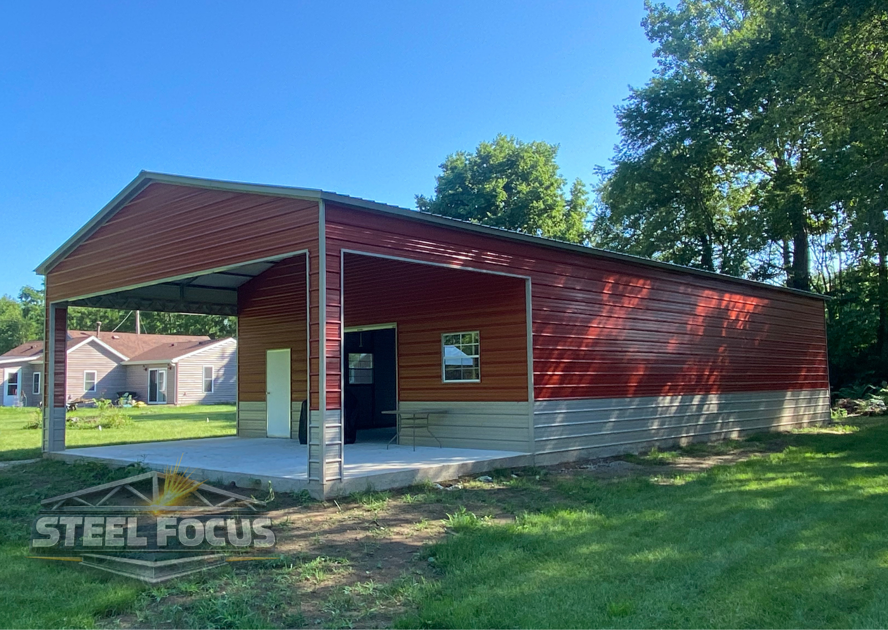 A redmetal building with a covered open front area, a small window, and a door on a concrete slab, located in a grassy area with trees and neighboring houses in the background.