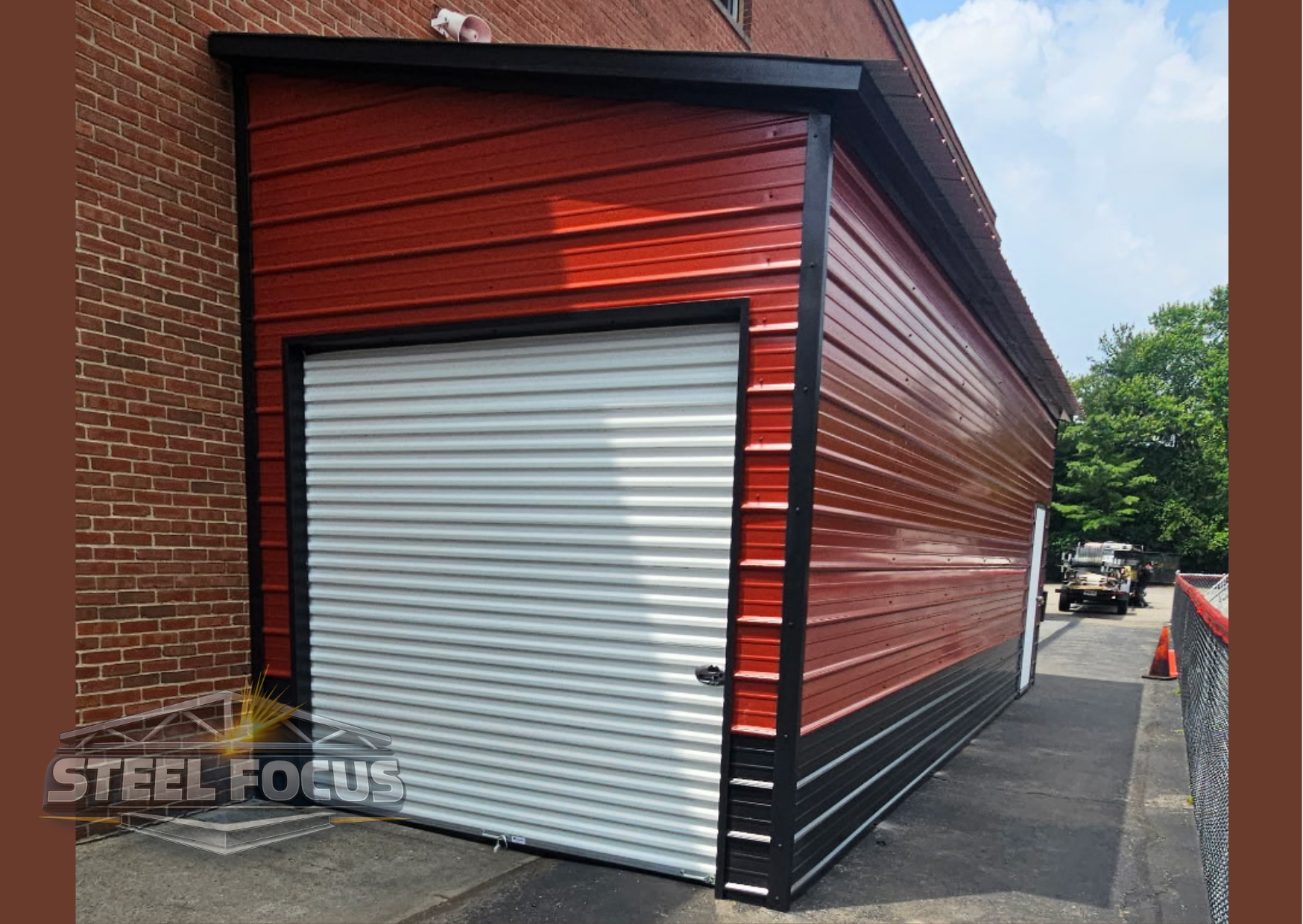 Red and black metal storage building with a white roll-up garage door, located next to a brick wall with a security camera on top. There is a trailer and orange traffic cone in the background, with green trees and a blue sky.