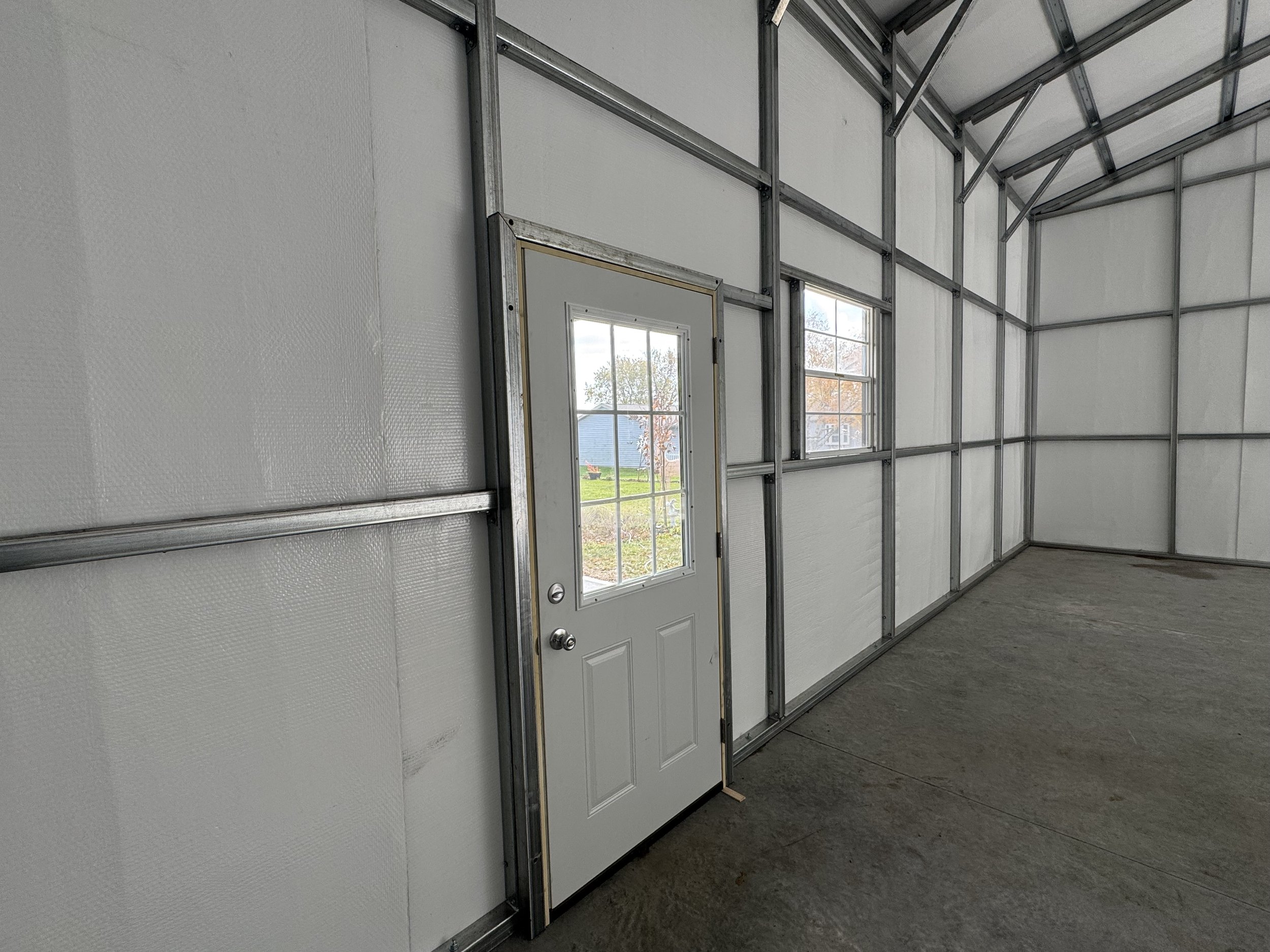 Interior of a partially constructed garage or building with metal framing, white insulated walls, an exterior door with glass panes, and two windows showing a yard with a blue house in the background.