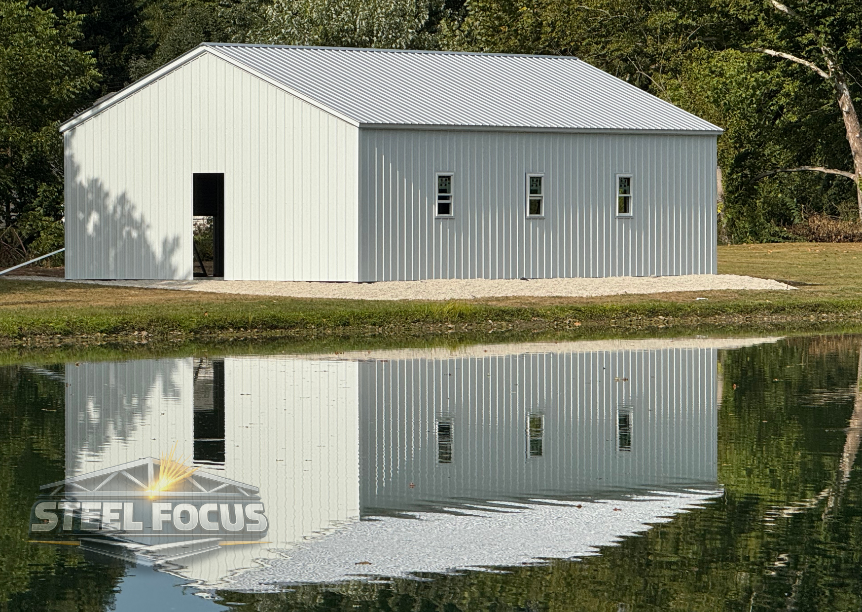 A white metal building with small windows and an open door, reflecting in a nearby body of water, with trees in the background.