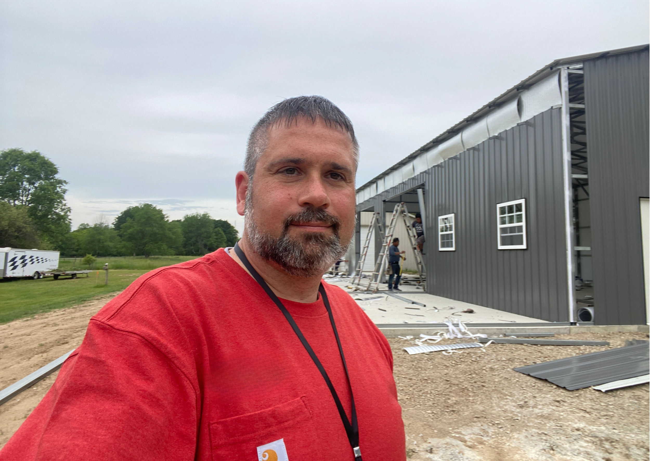 A man with gray hair and beard wearing a red shirt stands in front of a metal building under construction with workers on ladders working on the roof.