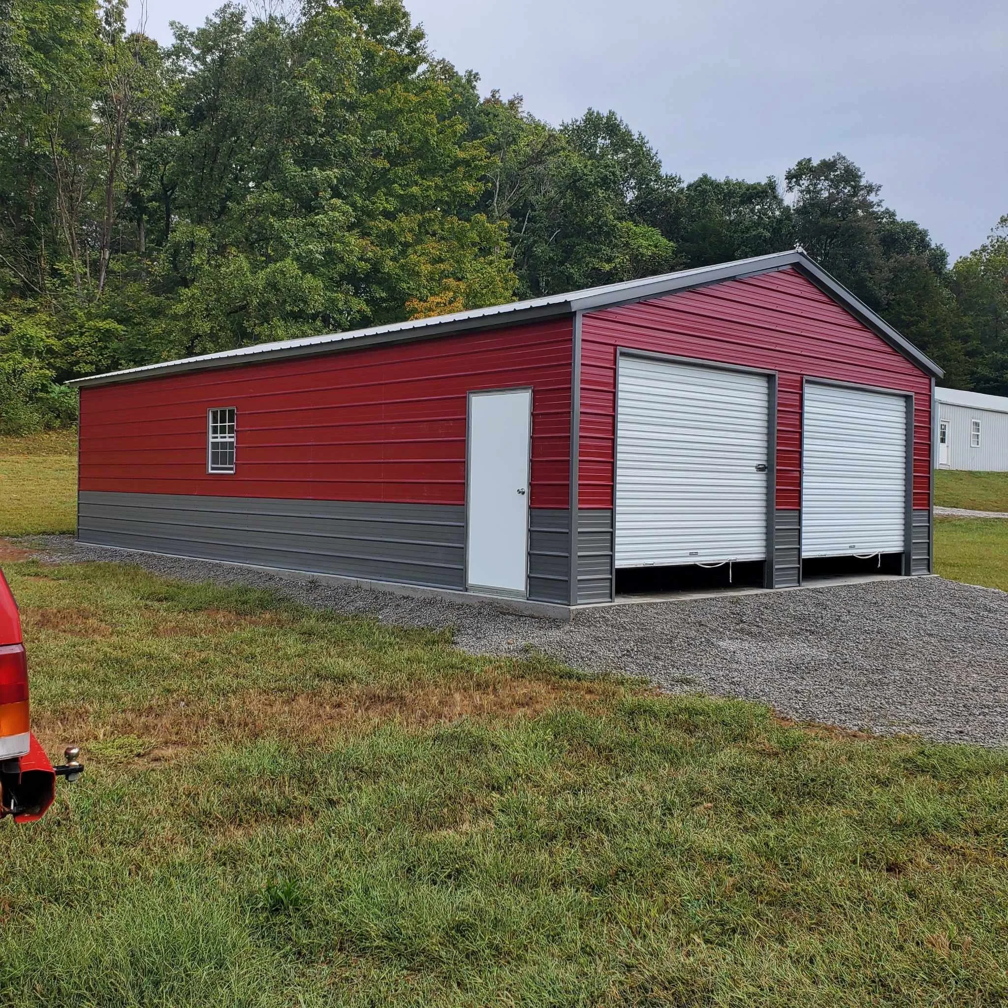 A red and gray metal storage building with two roll-up garage doors and a small side door, situated on a grassy field with trees in the background.