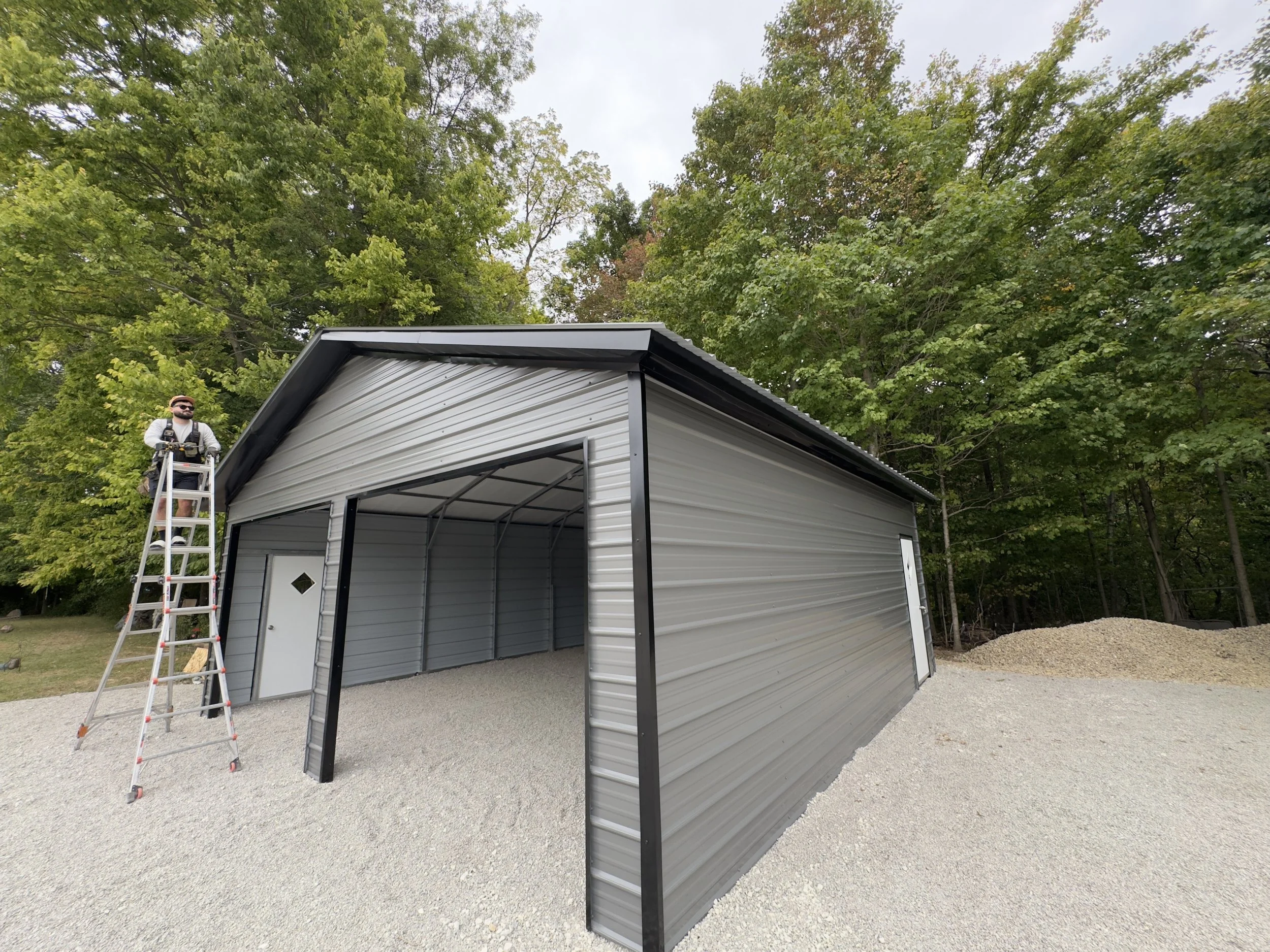 A person standing on a ladder next to a metal garage with a partially open front, surrounded by trees and gravel ground.