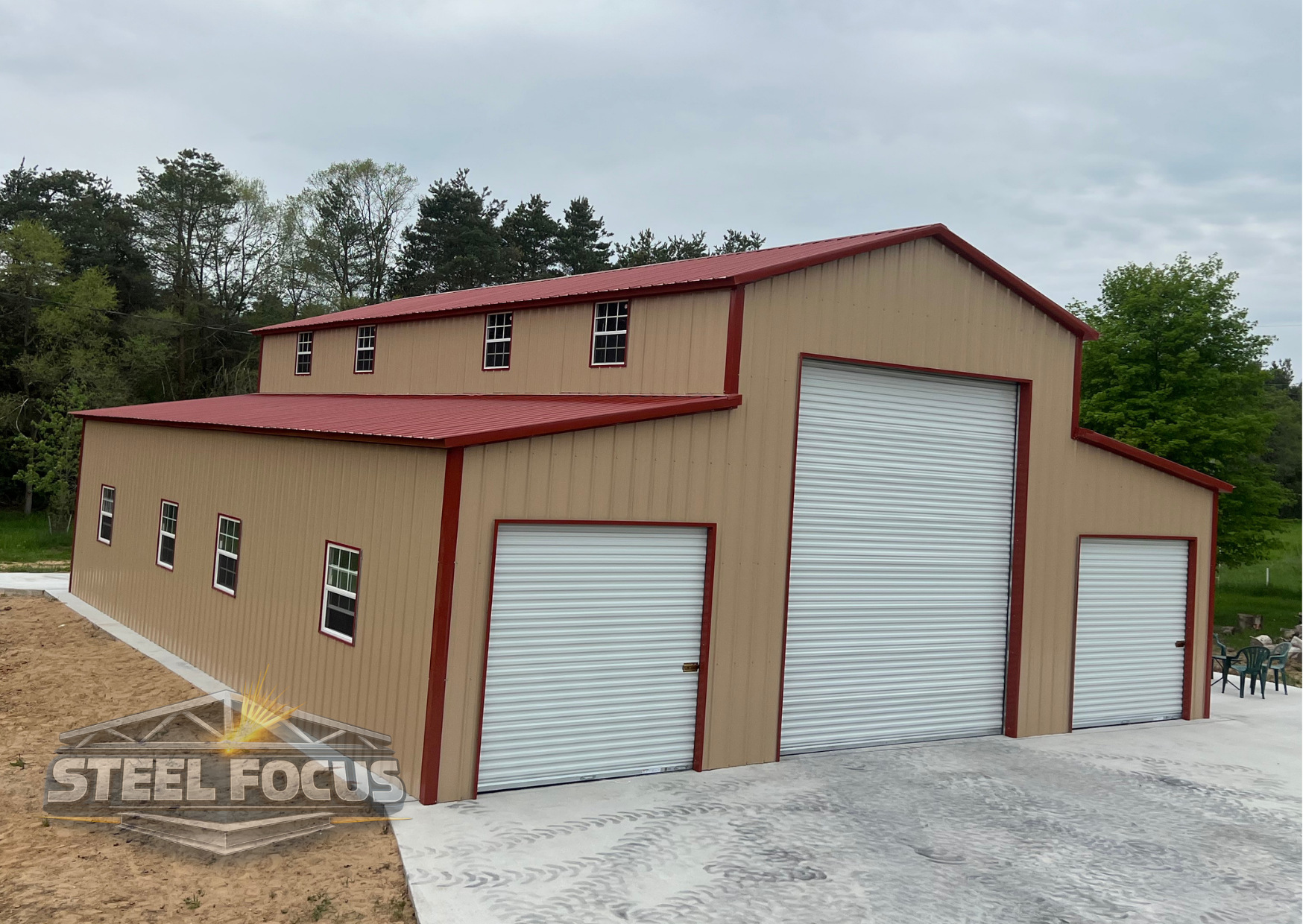 Large beige and red metal building with multiple roller doors, situated on a concrete and dirt surface, with trees in the background.
