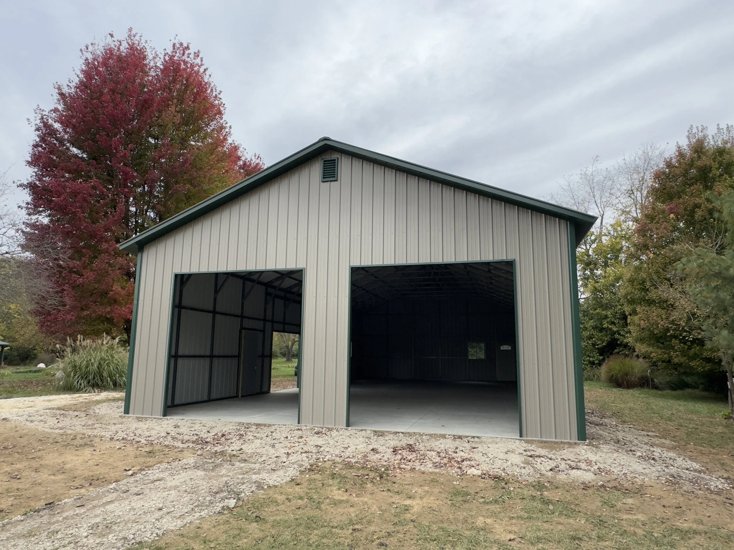 A large, empty metal framed garage or storage building with an open front, situated on a grassy area with trees in fall foliage in the background.