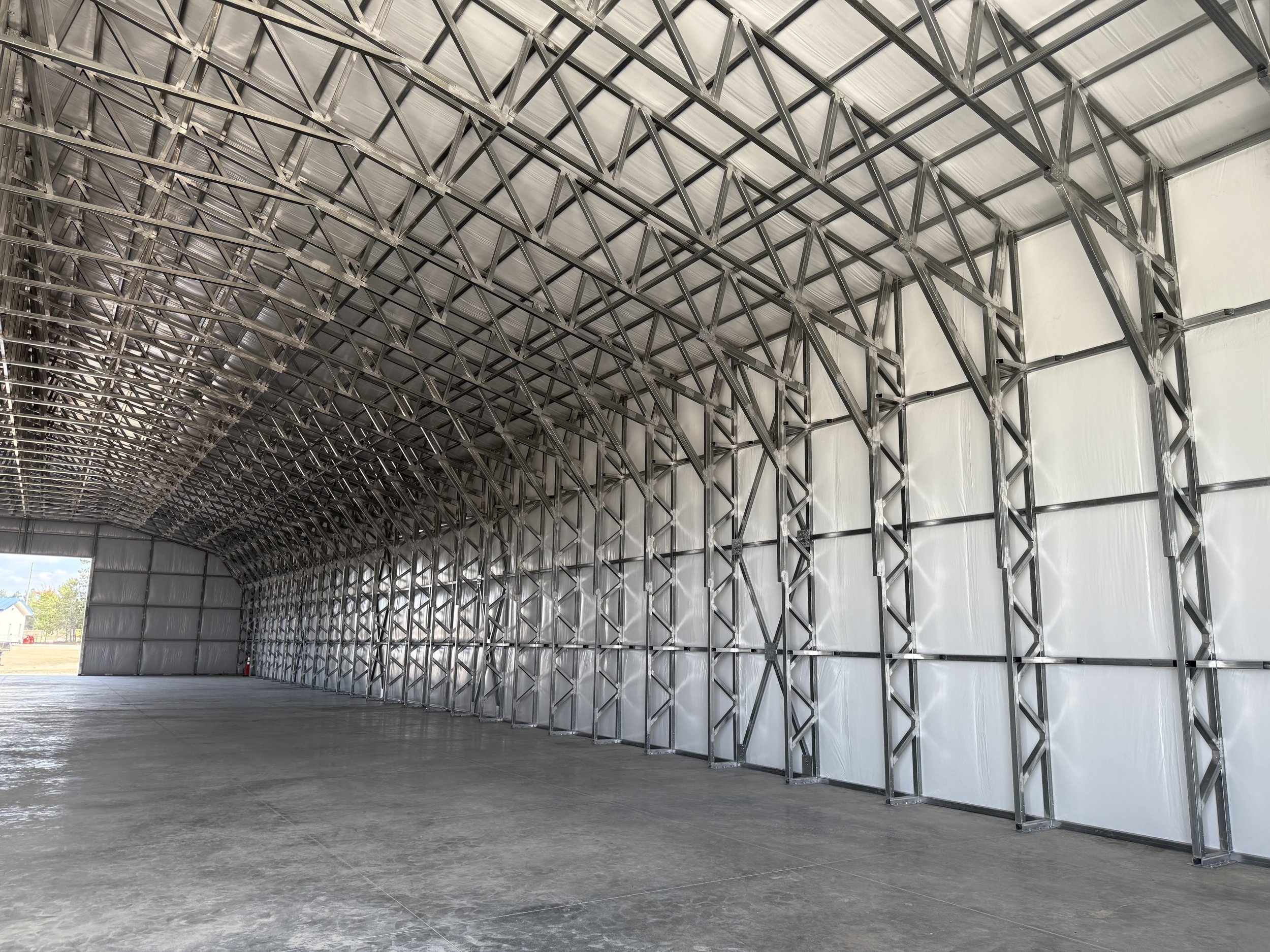 Interior of a large empty metal storage building or hangar with a high ceiling, metal trusses, and concrete floor, with an open door showing a view outside.
