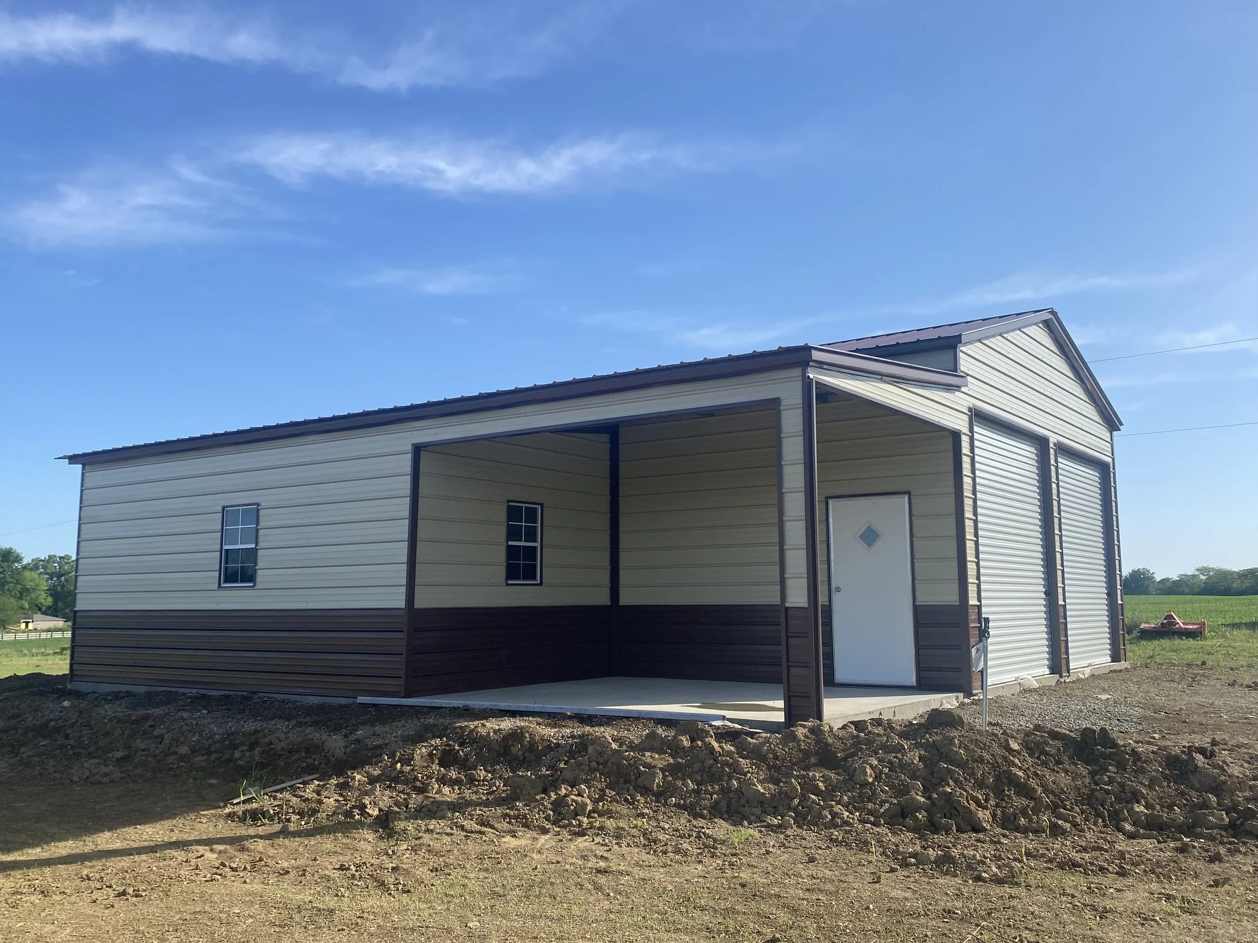 A newly constructed metal building with a covered porch, beige and brown exterior, located on a rural property with dirt ground and open fields in the background under a clear blue sky.