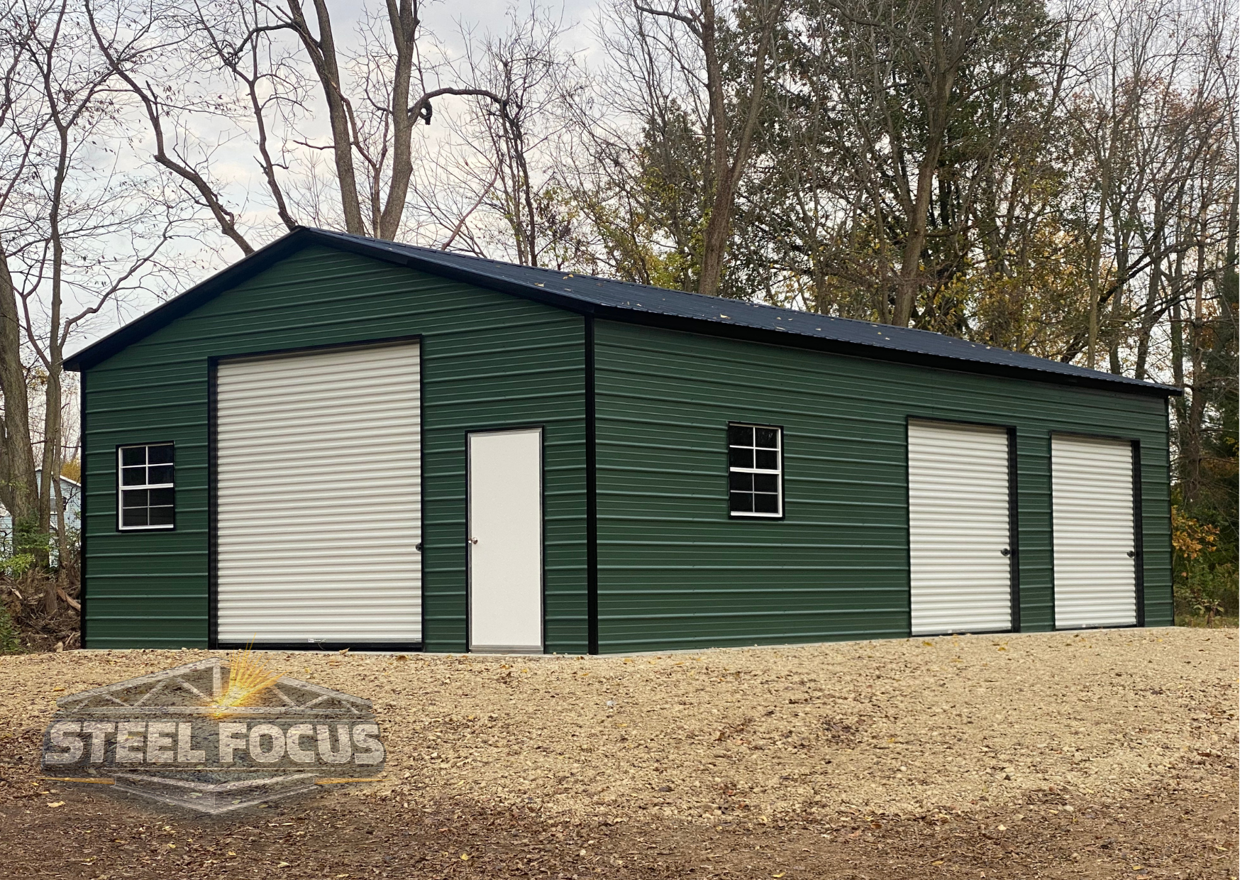 A large green metal building with three roll-up garage doors and a side door, set on a gravel surface with trees in the background.