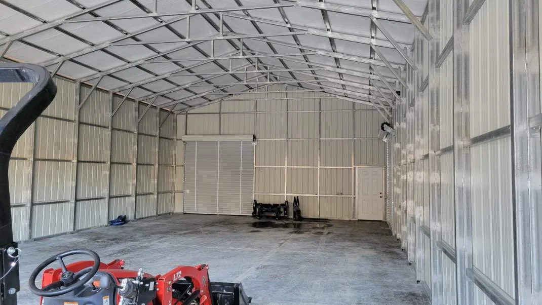 Inside a large metal storage building with a curved roof, concrete floor, and a roll-up door at the back. There is a small red piece of equipment in the foreground and some tools or equipment near the back wall.