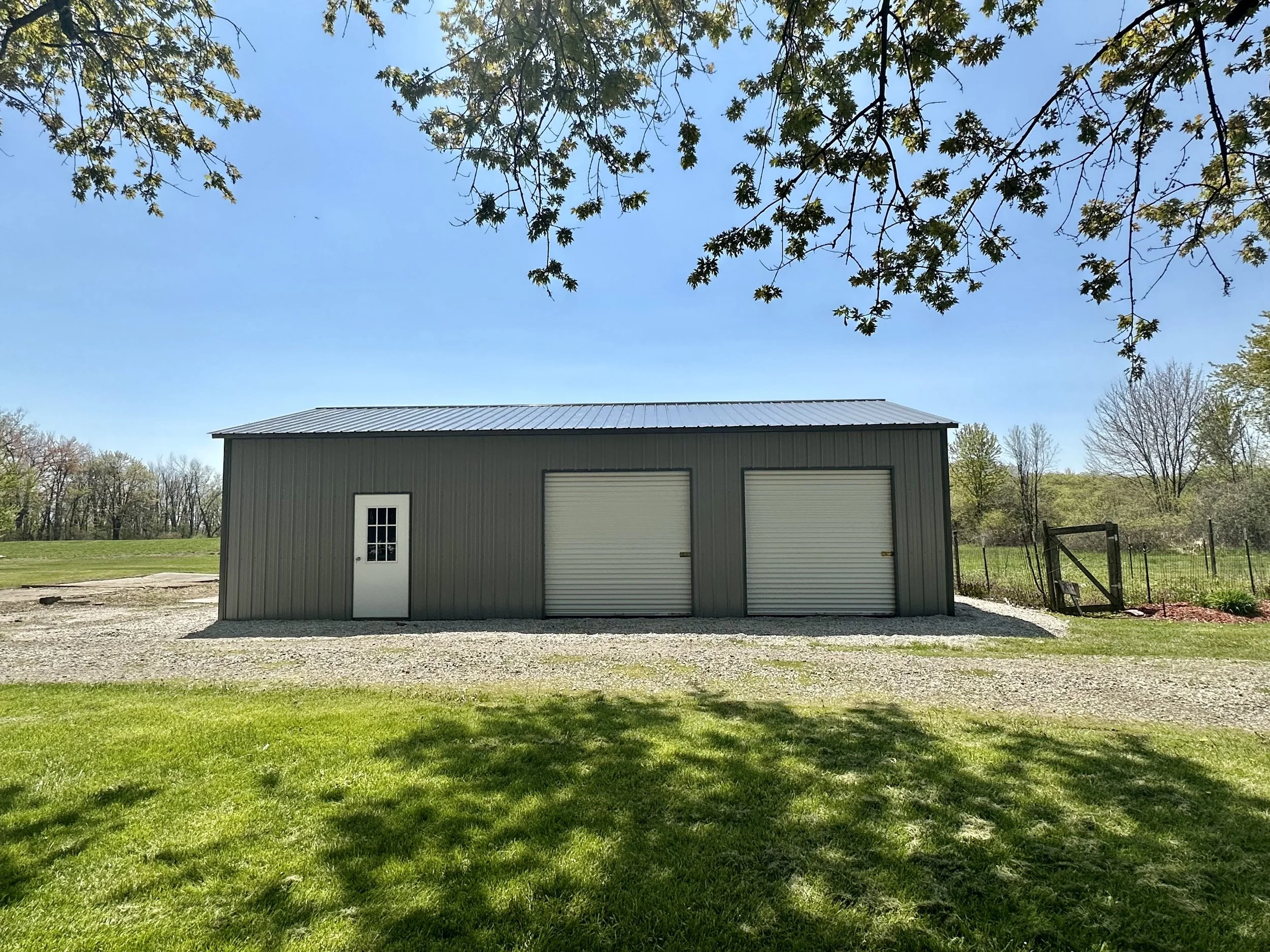 A small detached garage with two roll-up doors and a side door, surrounded by grass and trees under a clear blue sky.