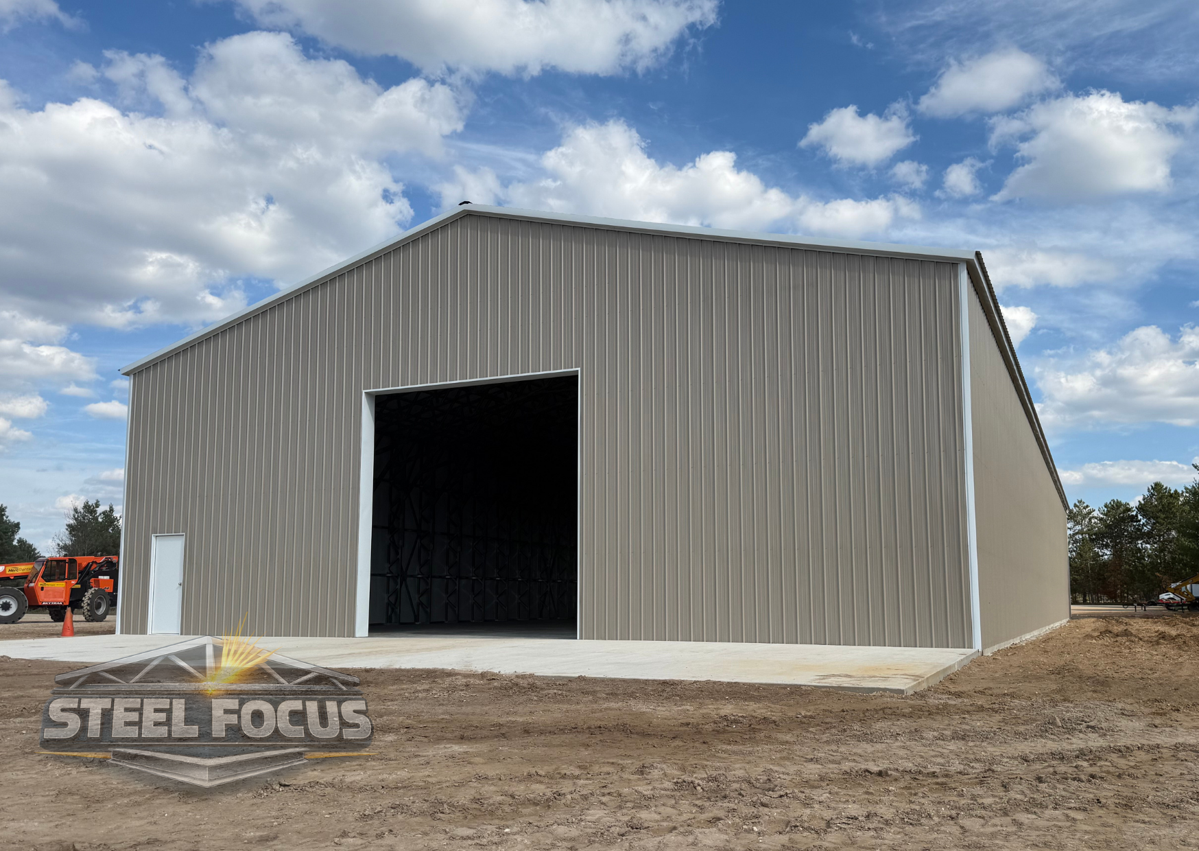Large metal storage building with a concrete driveway in front, under a partly cloudy sky, with construction equipment in the background.