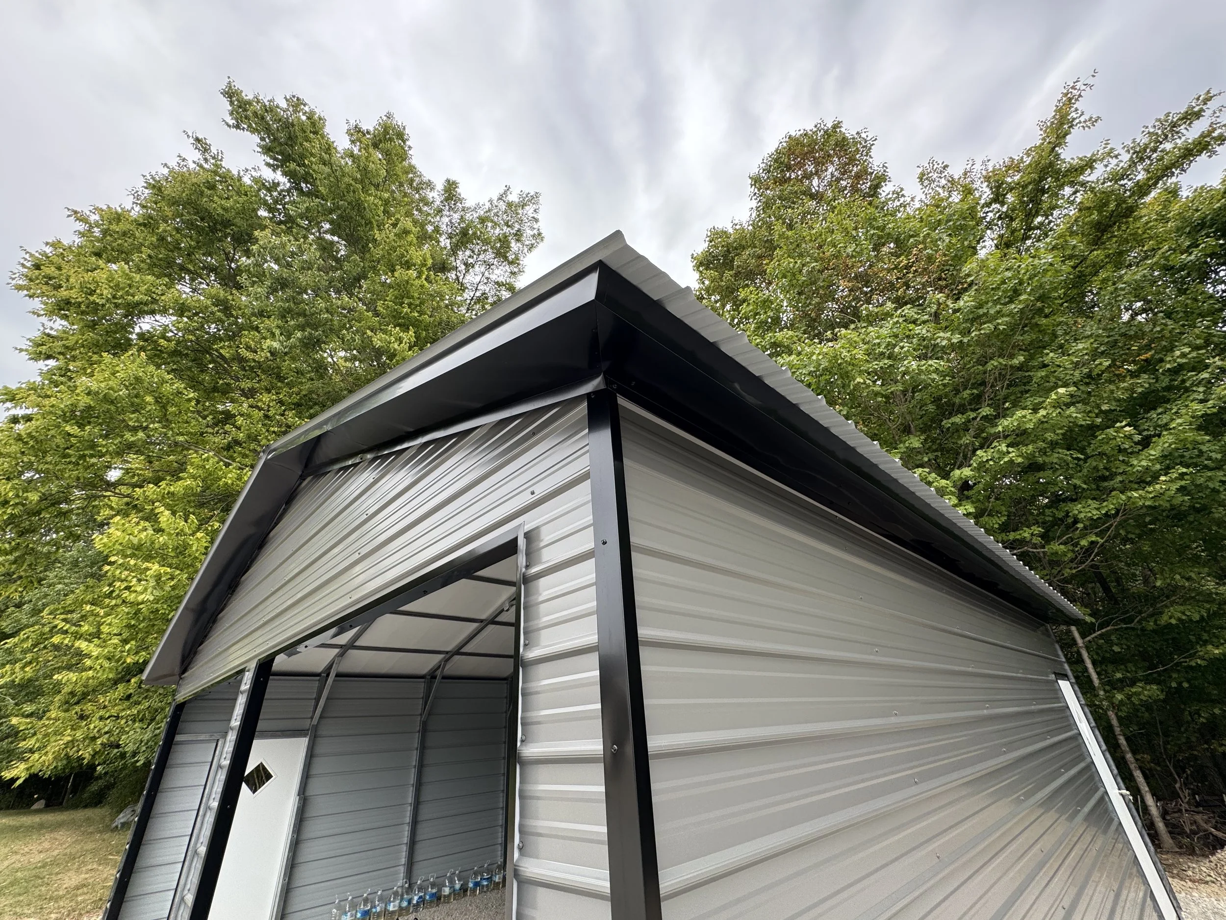 A metal storage shed with a curved white roof and side walls, set against green trees and an overcast sky.