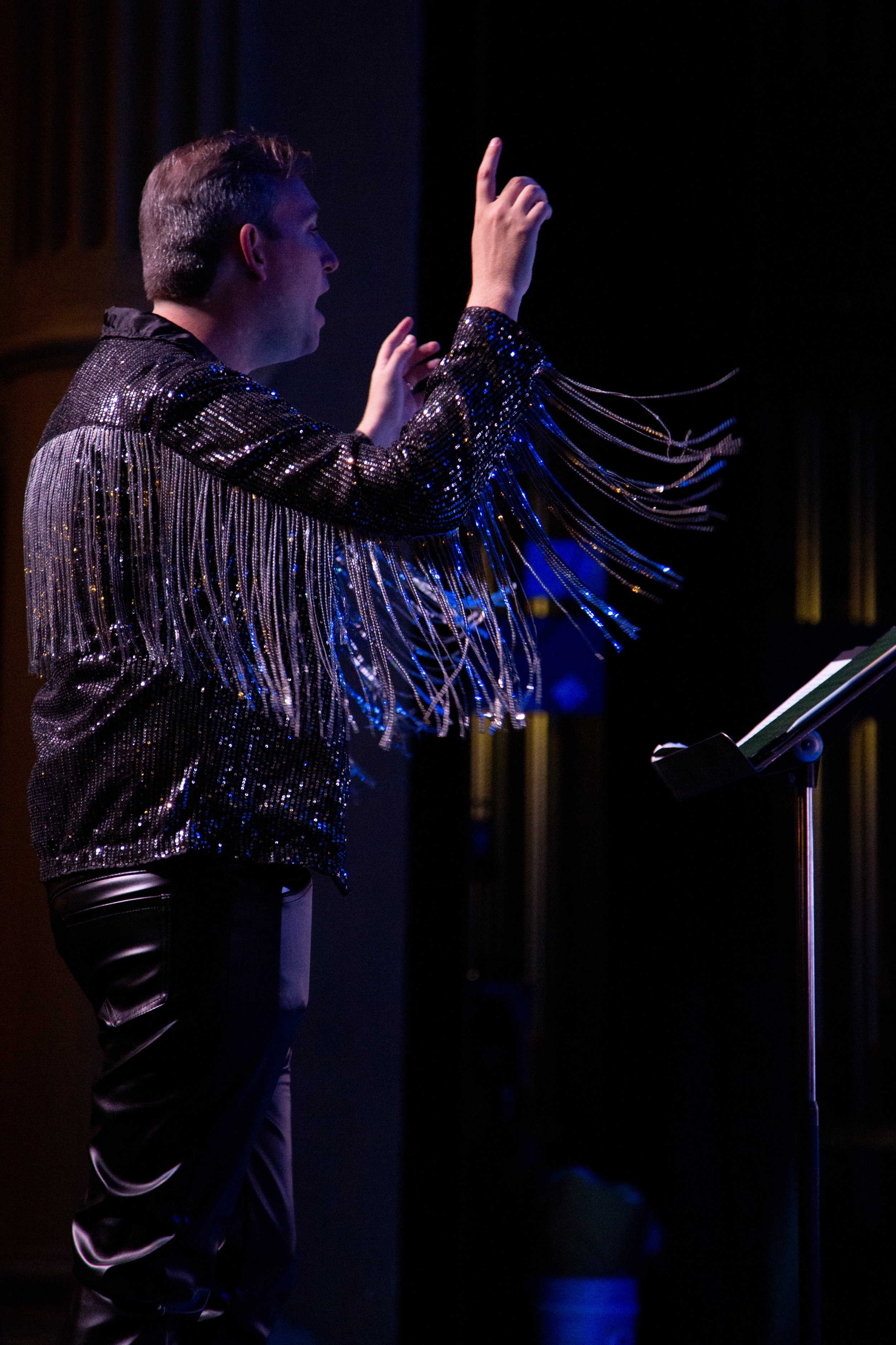 Performer on stage wearing a sparkly black jacket with silver fringe, gesturing with one hand raised, standing near a music stand in a dimly lit venue.
