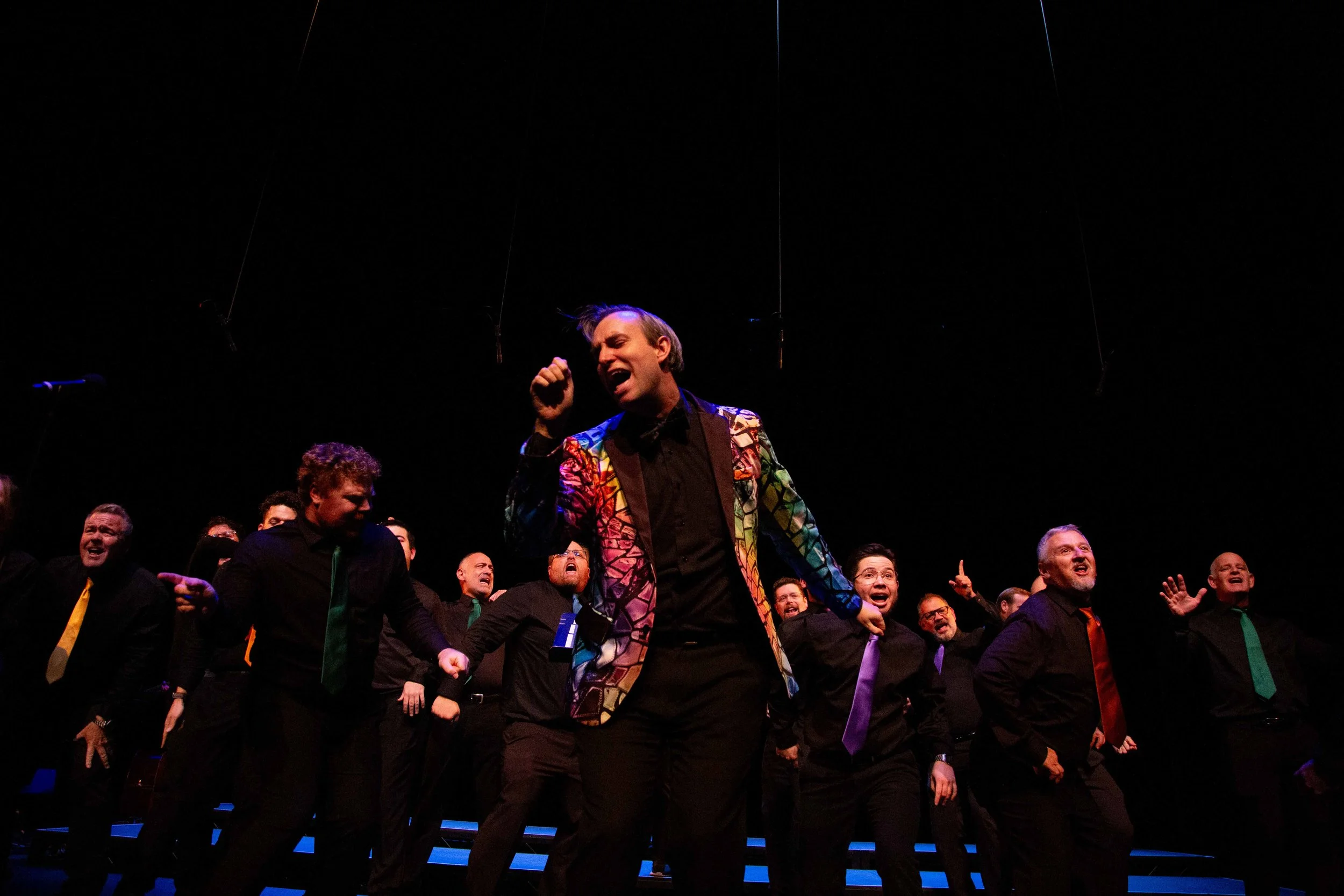 A man in a colorful, patterned blazer passionately singing or speaking to a crowd, surrounded by people dressed in black and colorful ties, all appearing engaged and enthusiastic.