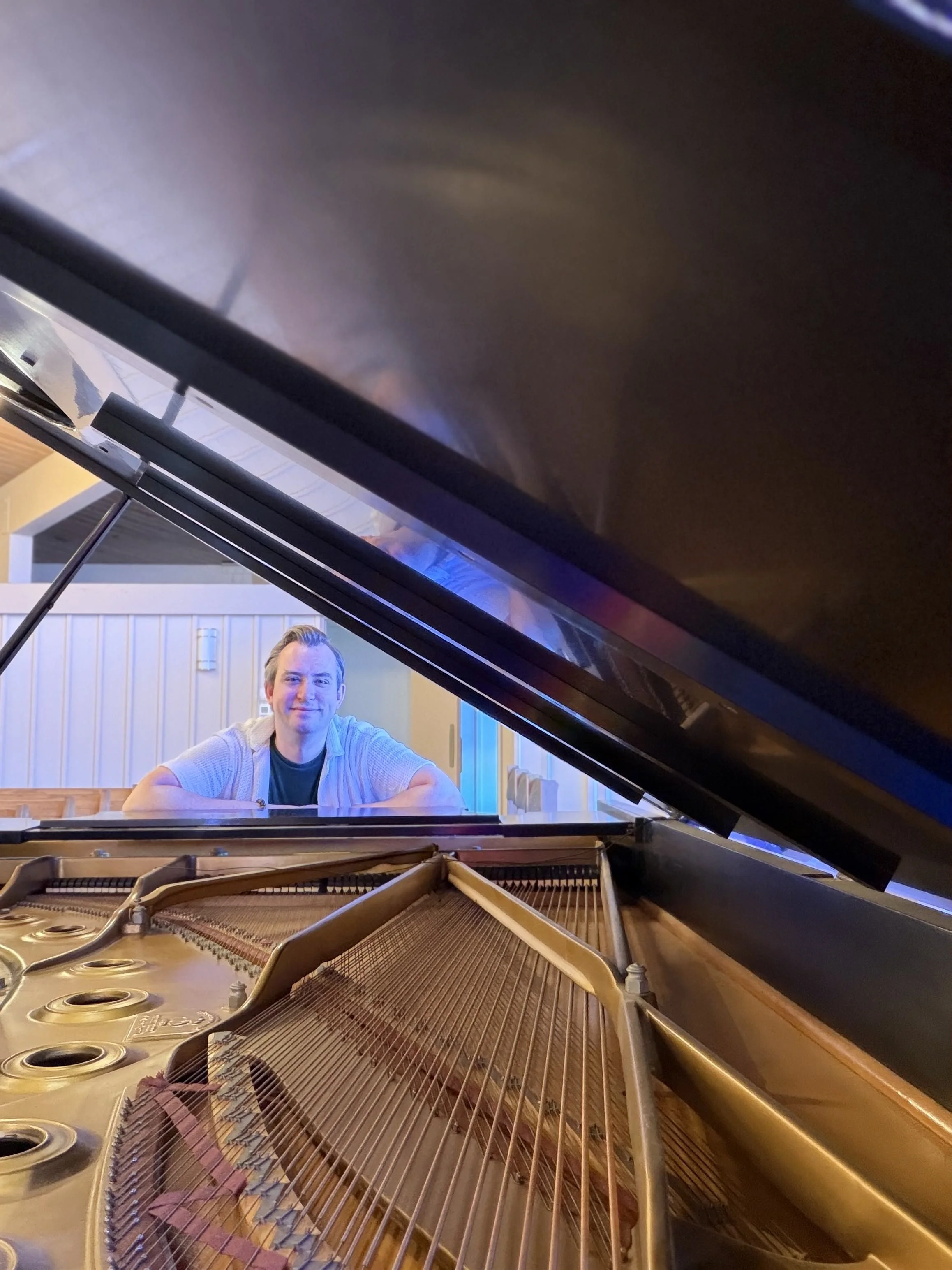 Man sitting at a grand piano with its lid open, showing the strings and internal components.