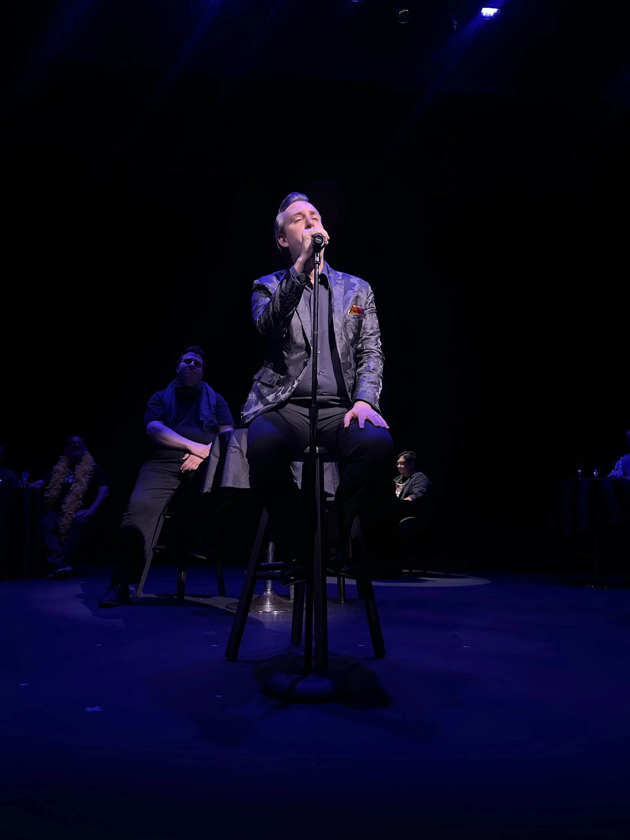 Man singing into a microphone on stage with purple stage lights, sitting in front of a chair, audience members watching.