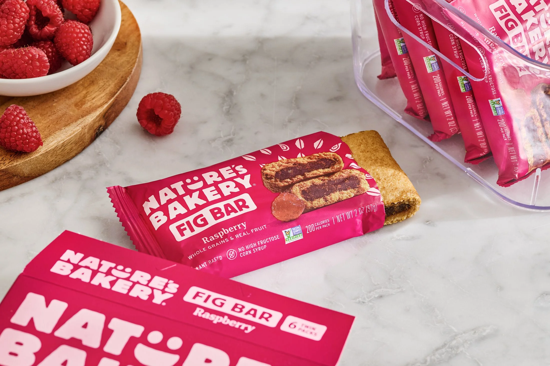 A close-up of a package of Nature's Bakery fig bars in raspberry flavor on a marble countertop, with fresh raspberries in a white bowl on a wooden tray in the background.
