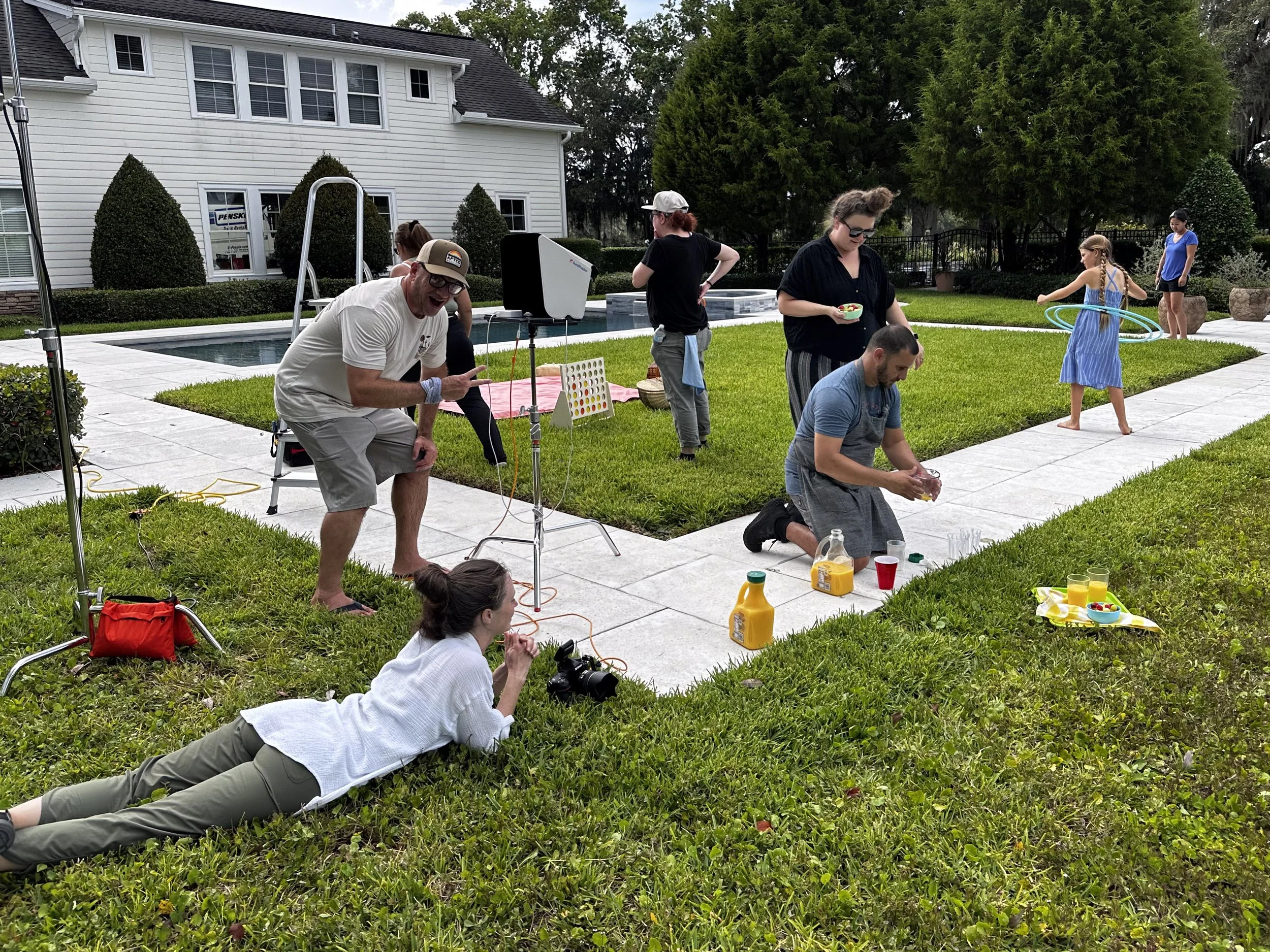 A group of people, including children and adults, are gathered outdoors in a backyard with green grass and trees, participating in a daytime photoshoot or filming session. Some are sitting or kneeling, and others are standing, with equipment and props around them near a pool and a white house.