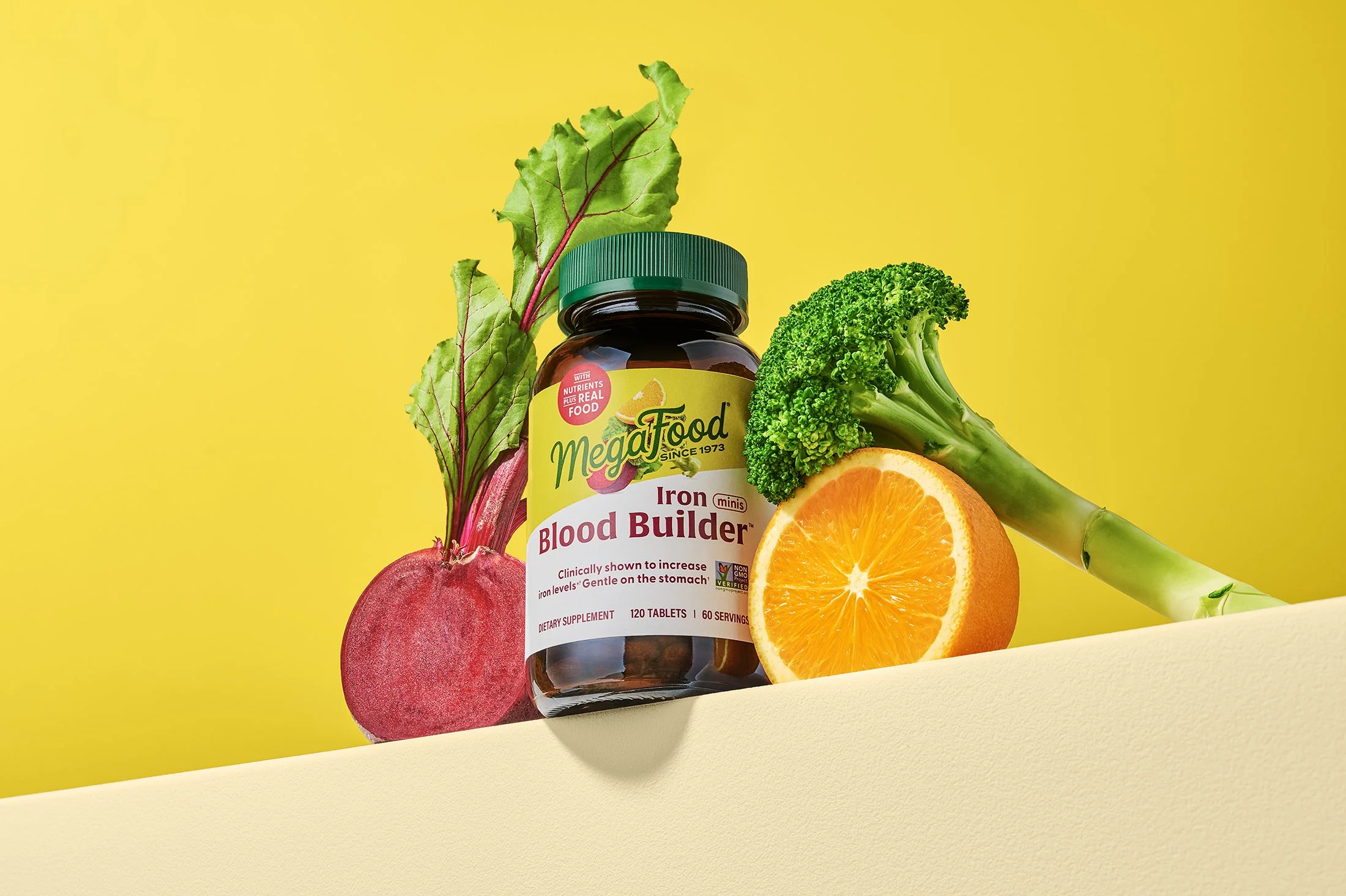 Bottle of MegaFood Blood Builder supplements surrounded by fresh vegetables including beets, broccoli, orange, and leafy greens against a vibrant yellow background. Product photography.
