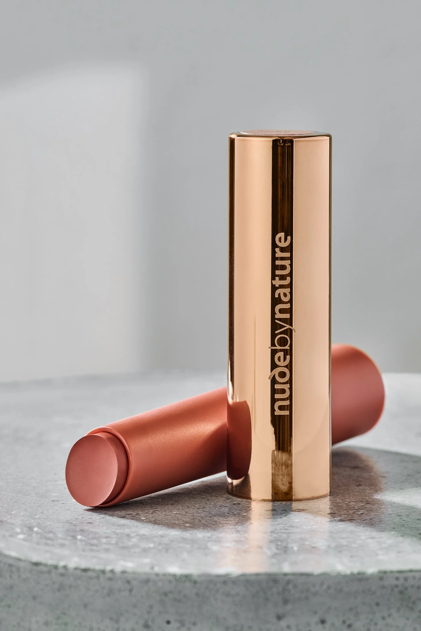 A person holding a rose gold lipstick with the brand name 'nude by nature' visible, and a pink lipstick tube is lying on a gray surface in the background.