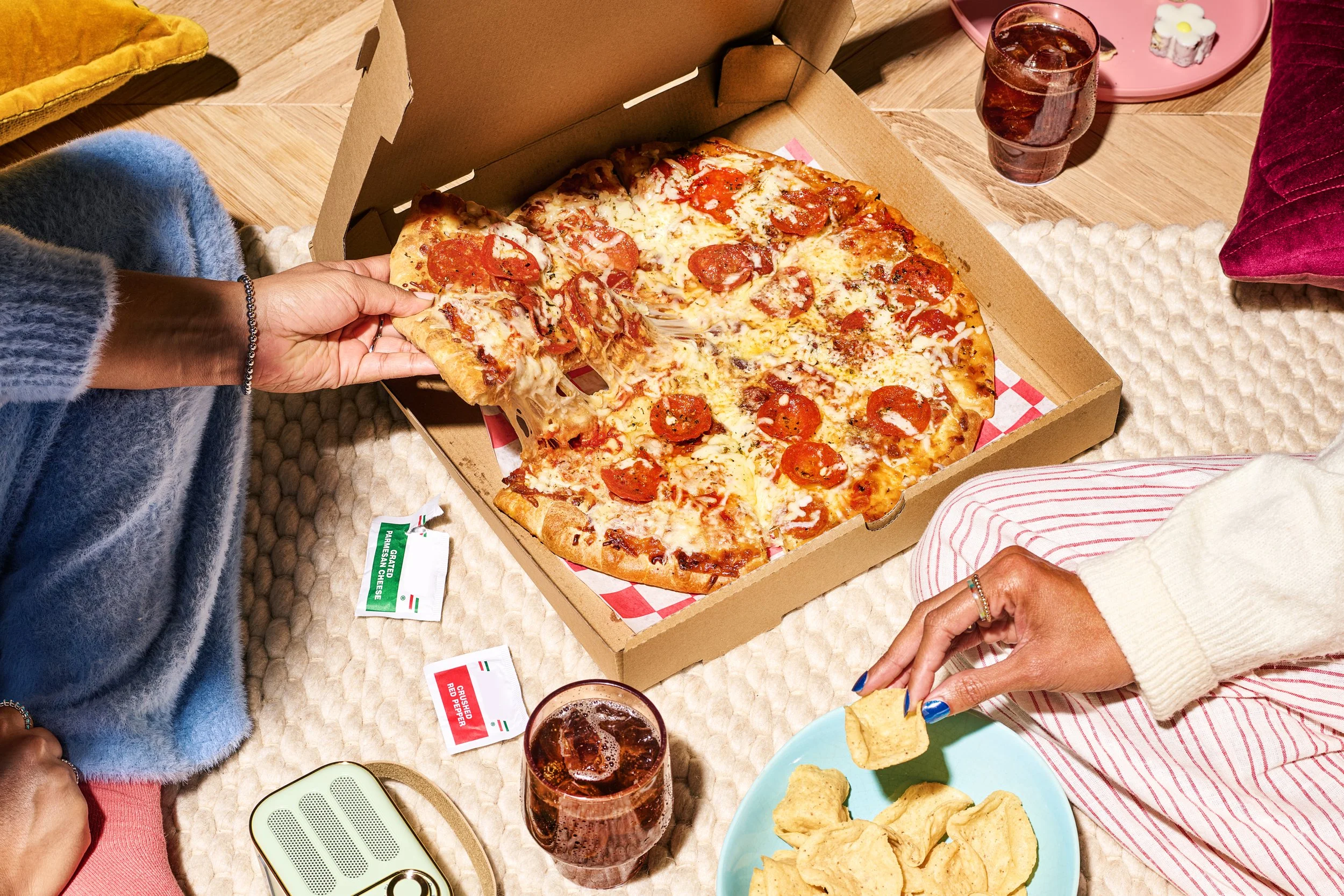 A pizza in a cardboard box with one slice being taken out, two glasses of soda, a plate of tortilla chips, and people sitting around a table.