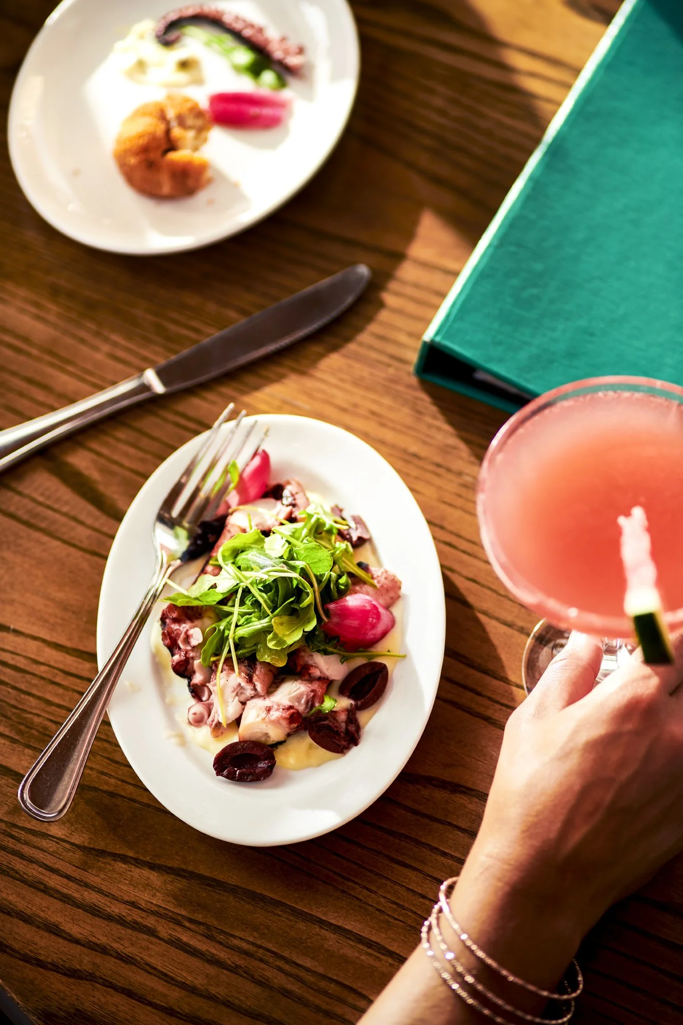 A person's hand with bracelets holding a pink cocktail glass at a wooden table. On the table, there's a white oval plate with seafood salad topped with mixed greens and radishes, and another white plate with assorted food. There is a green box and a 