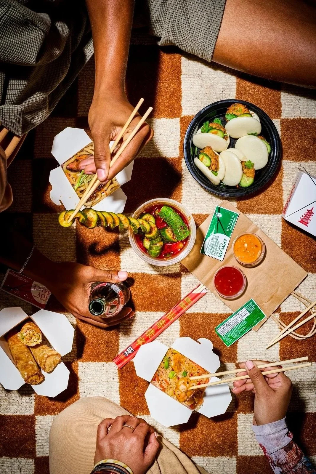 People having a meal with Asian cuisine, including spring rolls, sushi, and dipping sauces, on a checkered tablecloth.
