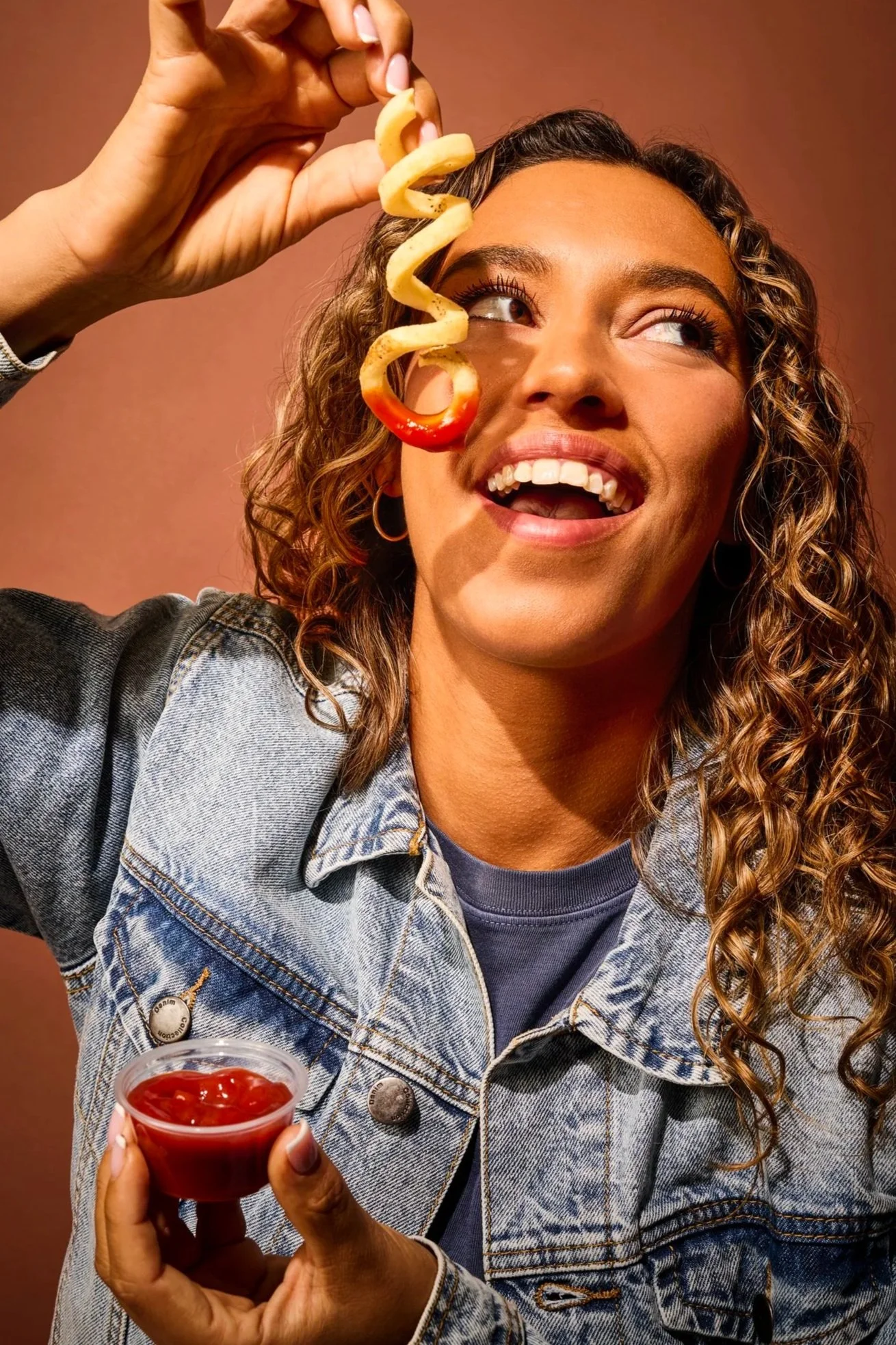 A woman with curly hair, wearing a denim jacket and earrings, holds a cup of ketchup and a curly fries snack.