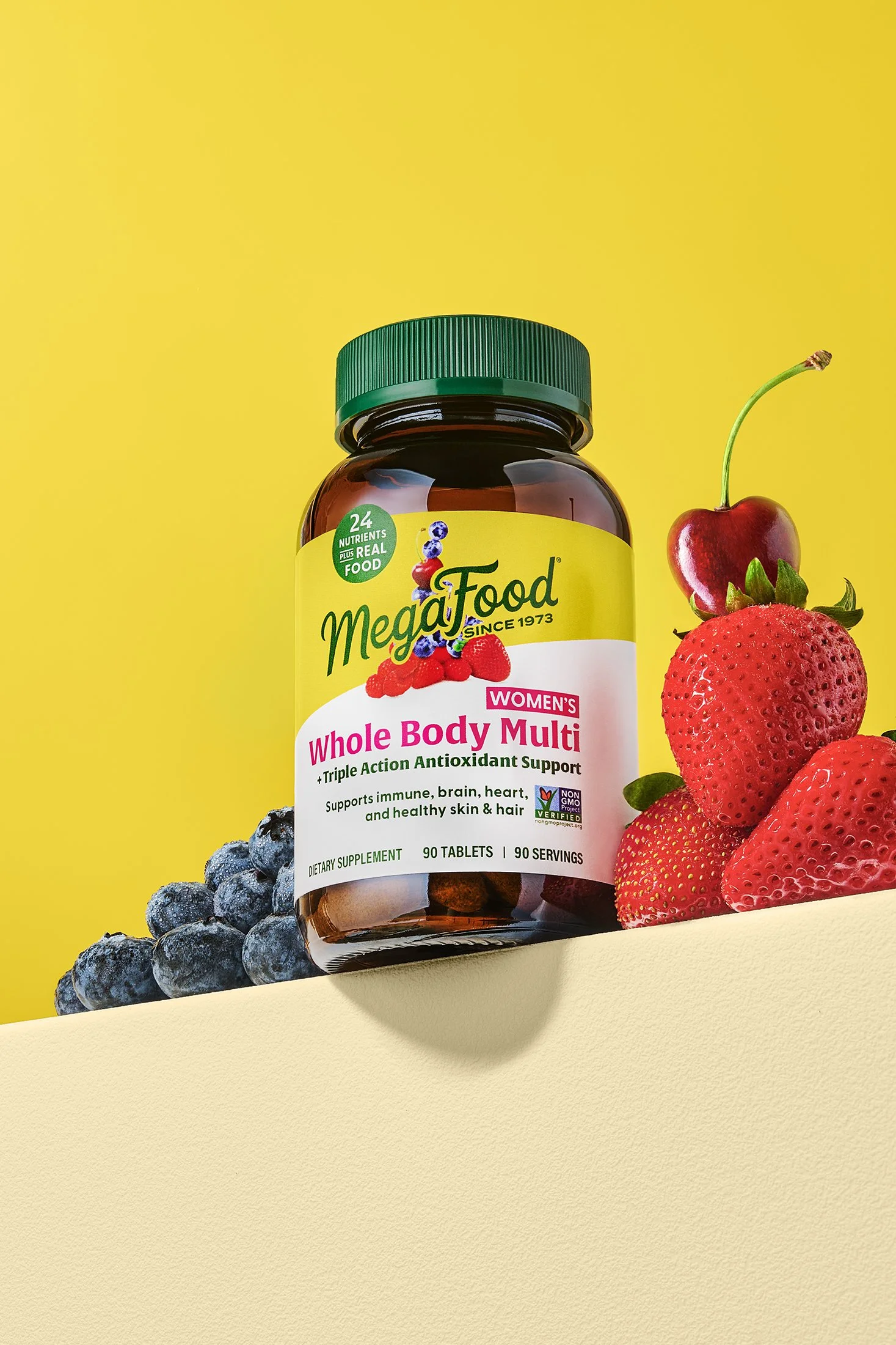 A bottle of MegaFood Whole Body Multi supplement with blueberry, raspberry, and strawberry fruits placed around it, against a yellow background.