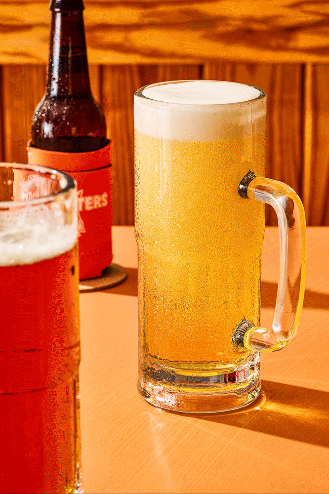 A glass beer mug filled with sparkling beer and foam on top, placed on a wooden table with a bottle of beer in a red koozie in the background.