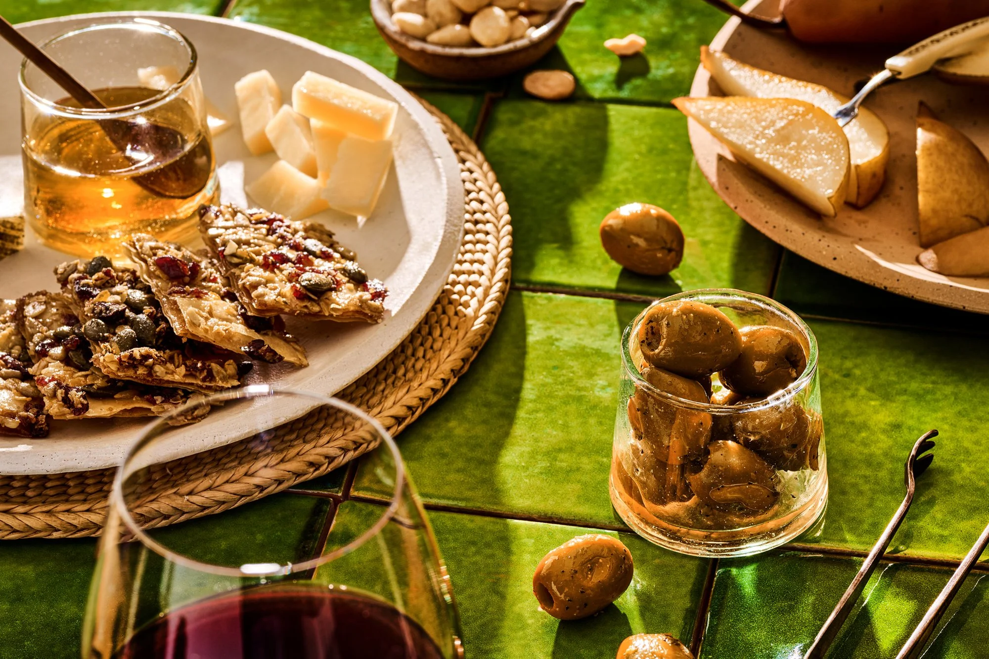 A table setting with cheese, nuts, dried fruit, and glasses of red wine and a drink with olives on a green tiled table.