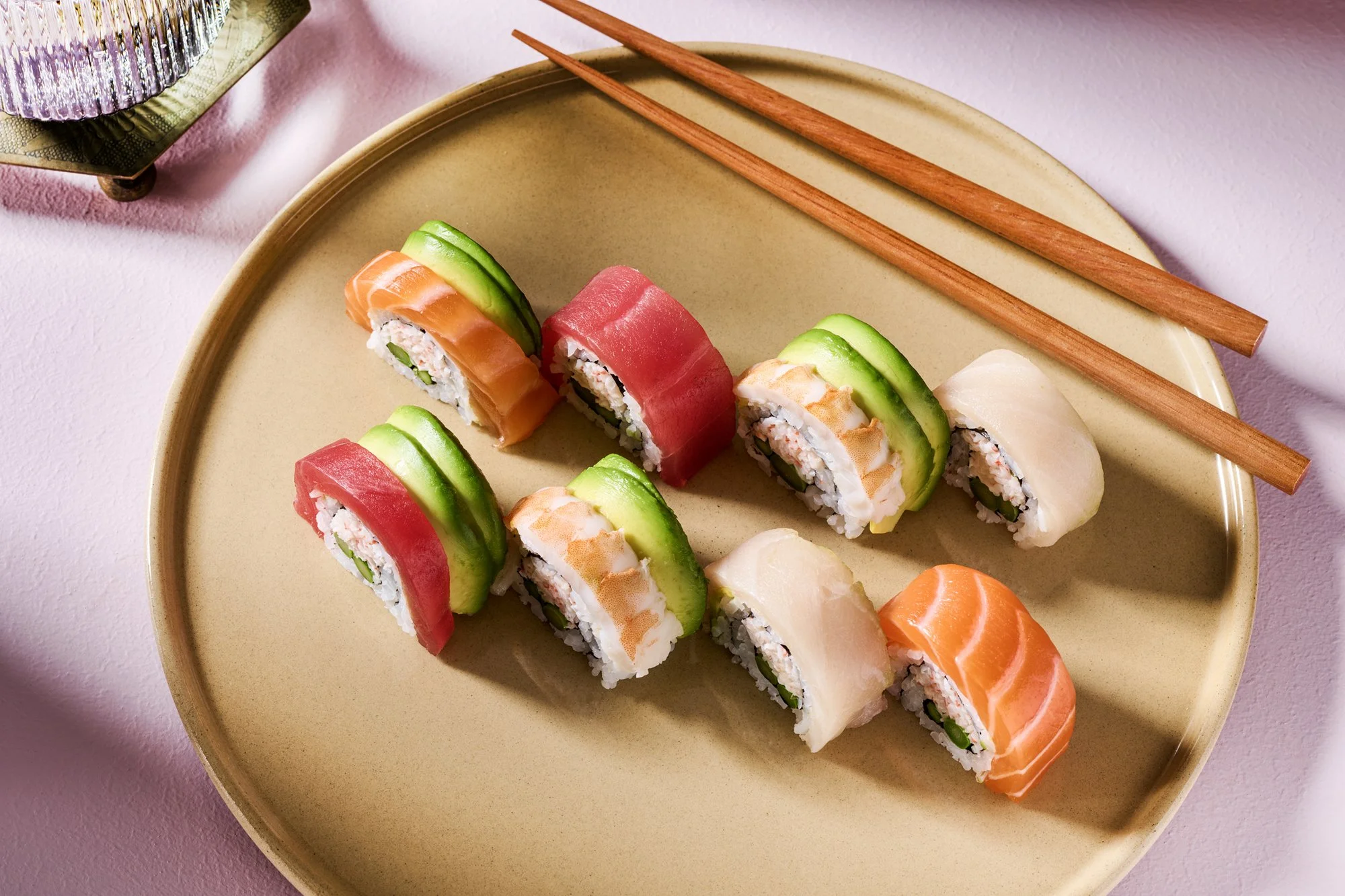 Sushi rolls with various fish and avocado on a beige plate, with wooden chopsticks resting on the side, placed on a pink surface.