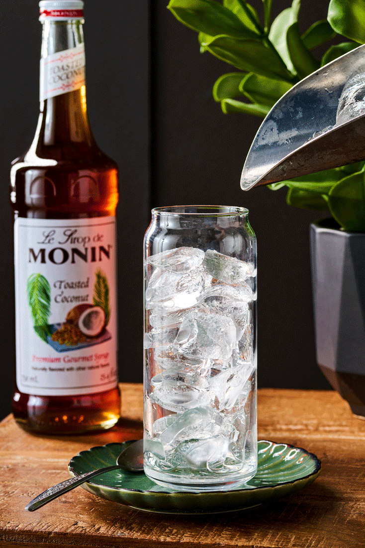 Cocktail photography gif of a glass filled with ice cubes on a green plate with a spoon, a bottle of Monin toasted coconut syrup in the background, and a plant in a gray pot.