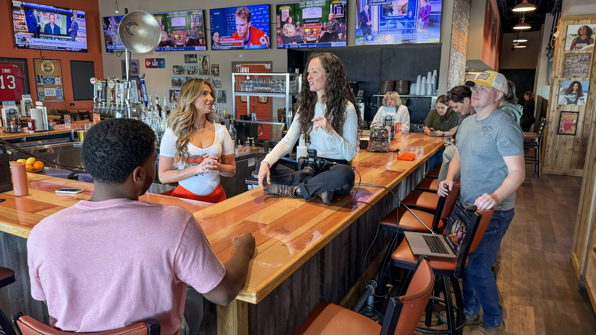 A group of people gathered inside a bar with multiple television screens showing sports and news. Two women are standing and talking by the bar counter, with one holding a camera. Several people are seated, some with drinks, while others are using their phones. The bar has various taps, posters, and signs, creating a lively atmosphere.