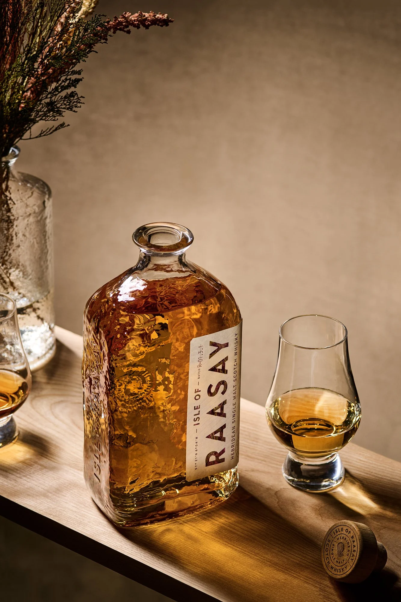 A bottle of red whisky with a white label, along with a glass of whisky on a wooden tray, surrounded by a silver vase with flowers and other glasses. CPG Product beverage photography Tampa Florida by Nutmeg Imageworks.