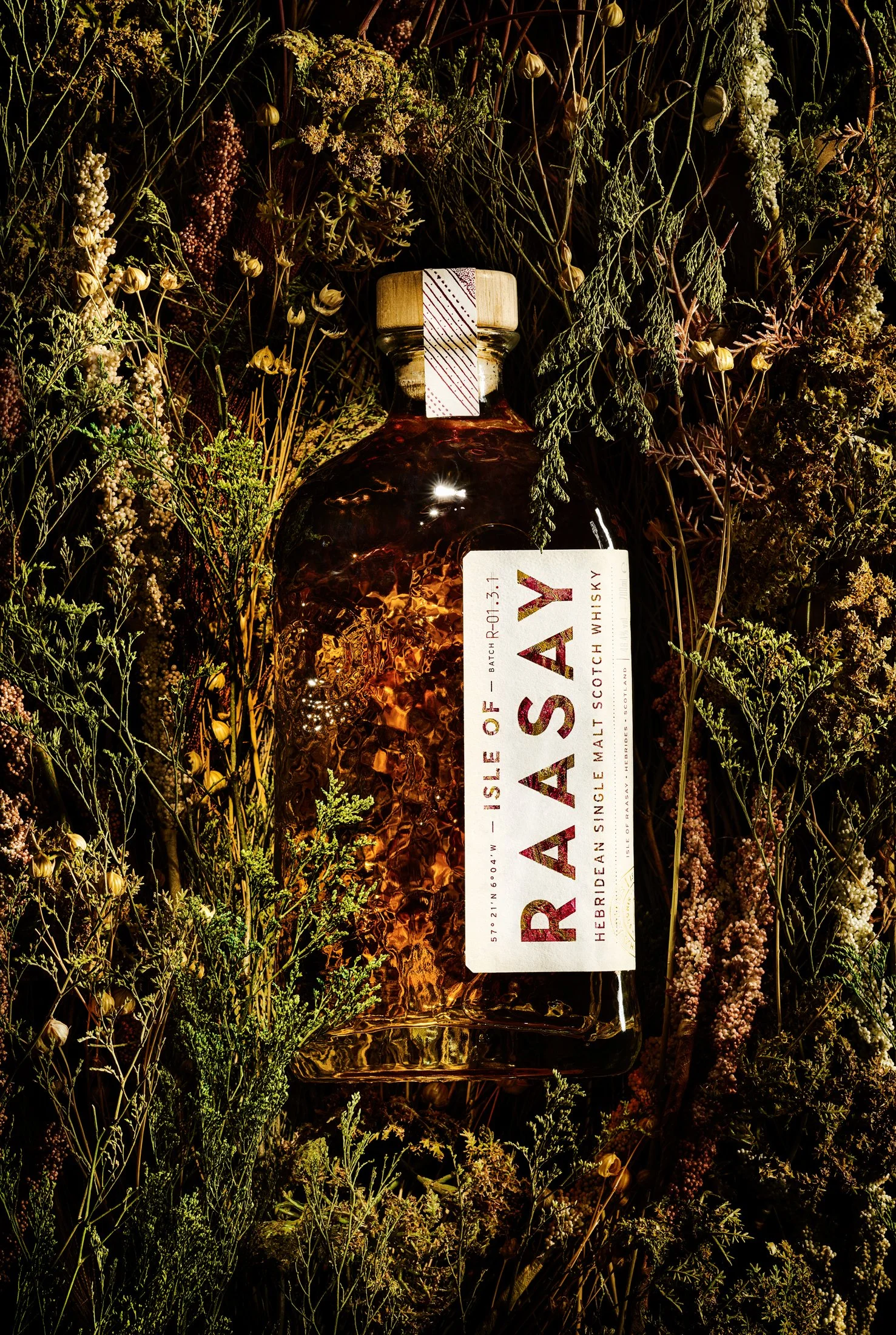 A bottle of single malt Scotch whisky is surrounded by green and brown dried plants and flowers, with warm lighting highlighting the bottle.