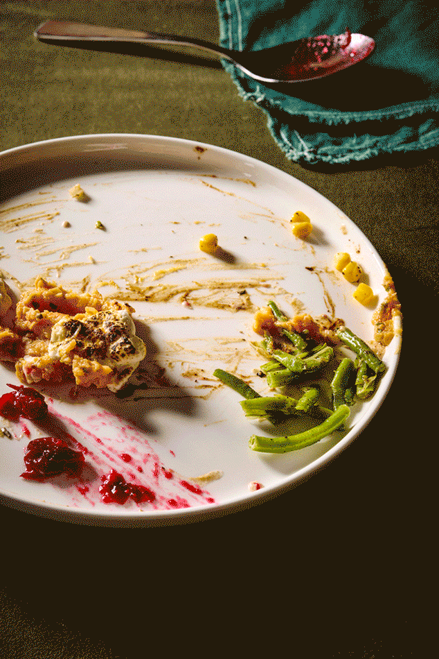 Empty white plate with leftover food and sauce, green vegetables, and a few yellow corn kernels, on a dark table with a spoon and a teal napkin nearby.
