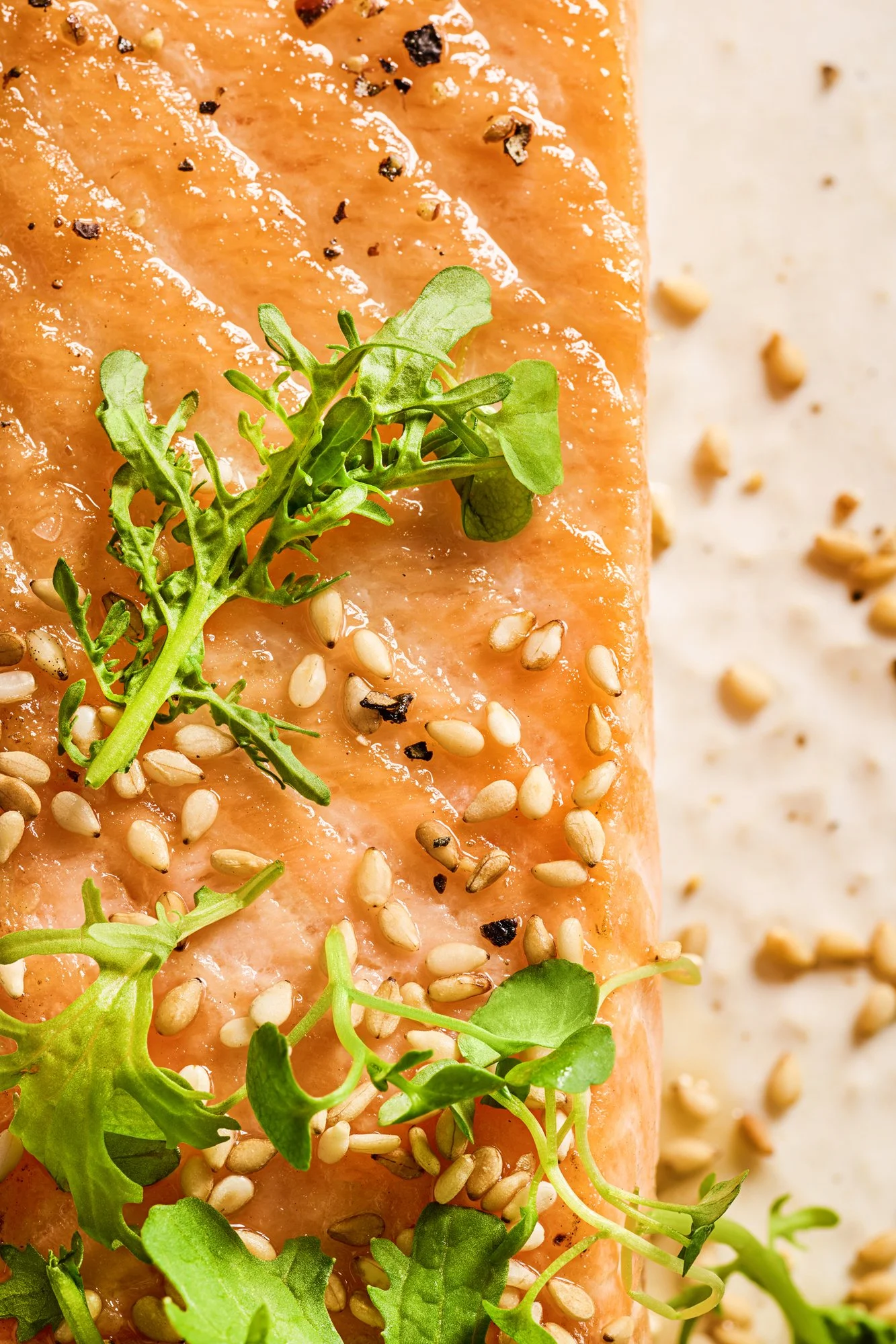 Close-up of a cooked salmon fillet garnished with fresh microgreens and sprinkled with sesame seeds and black pepper, on a white surface.