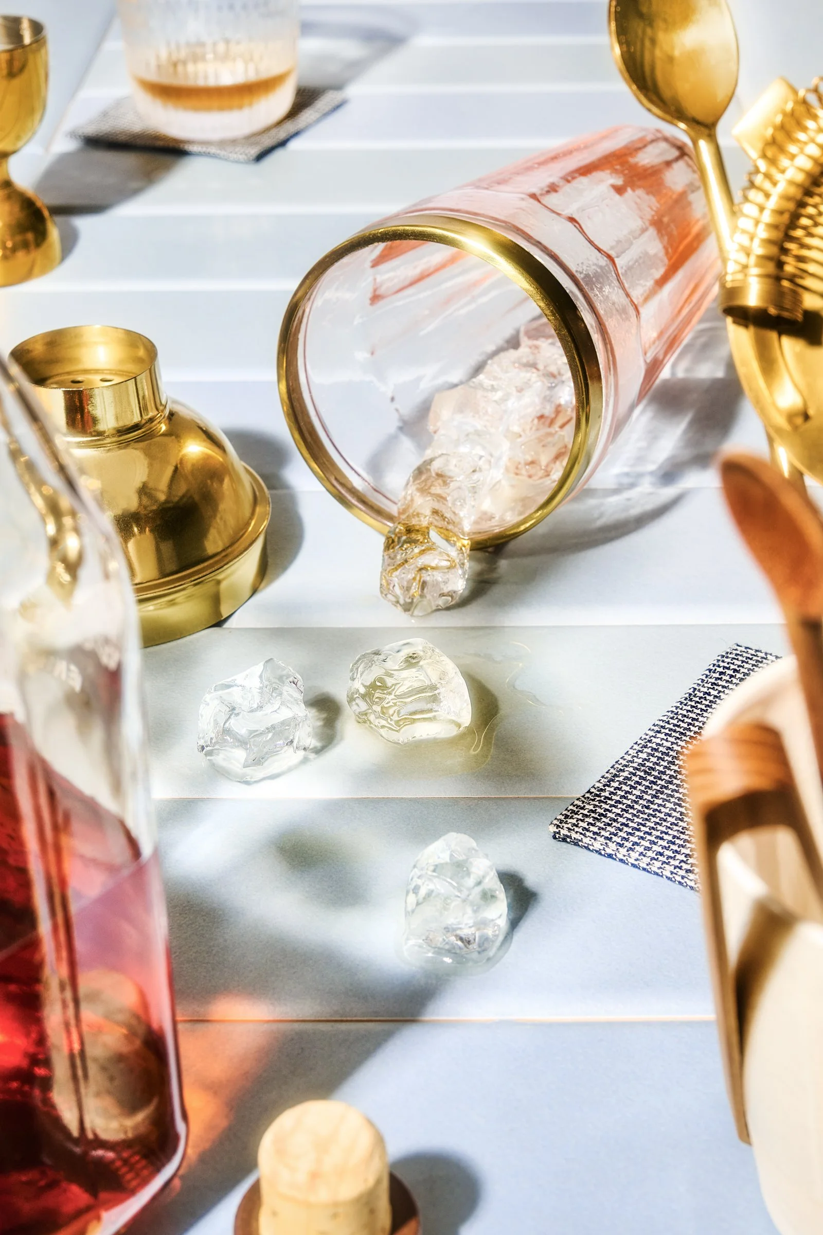 A table with a pink glass container tipped over spilling ice, surrounded by gold-colored candle holders and bottles, on a white wooden surface with a small piece of cloth nearby.