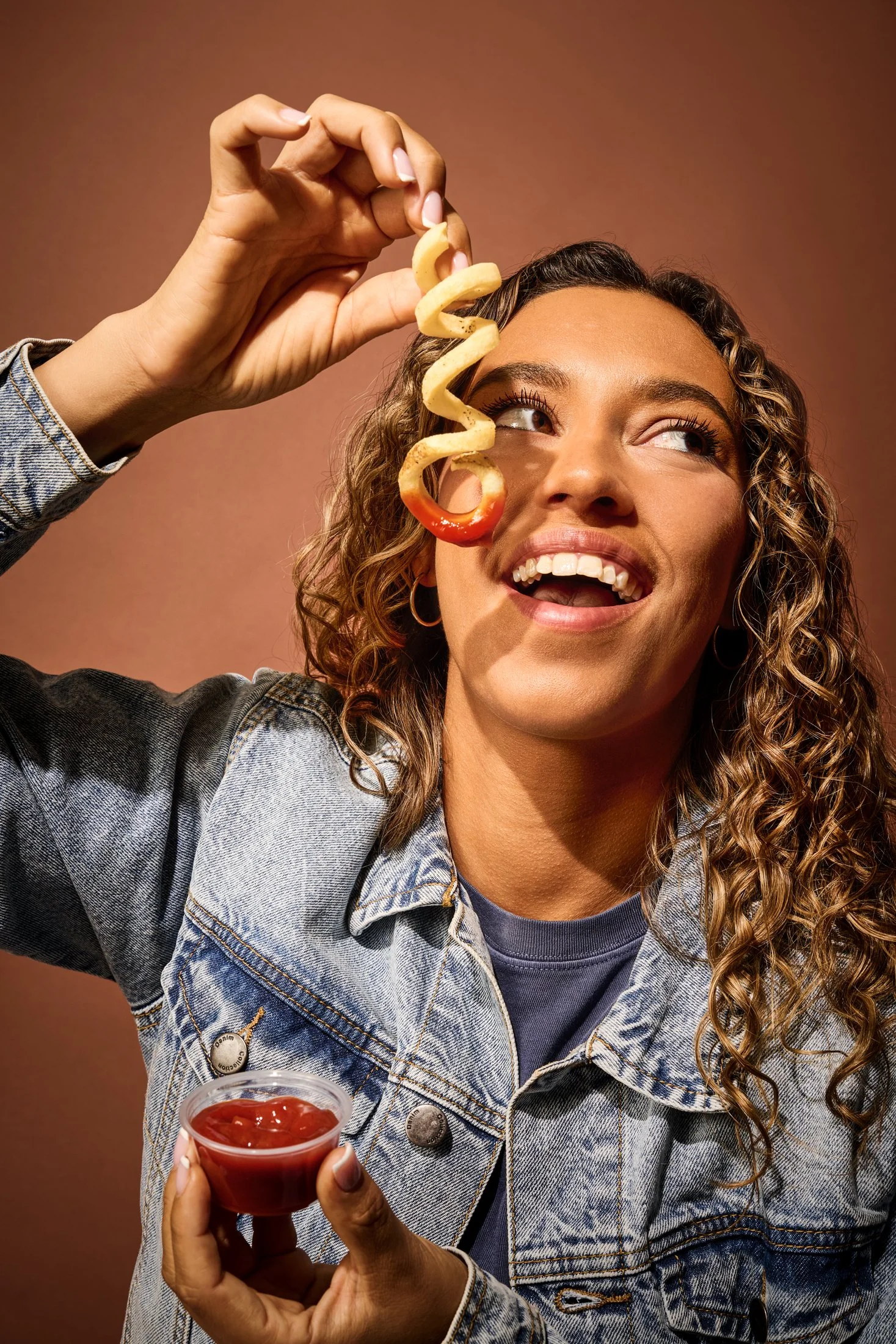 A woman with curly hair wearing a denim jacket, smiling as she holds a spiral-cut potato chip near her face and a small cup of ketchup. The background is a solid brown color.