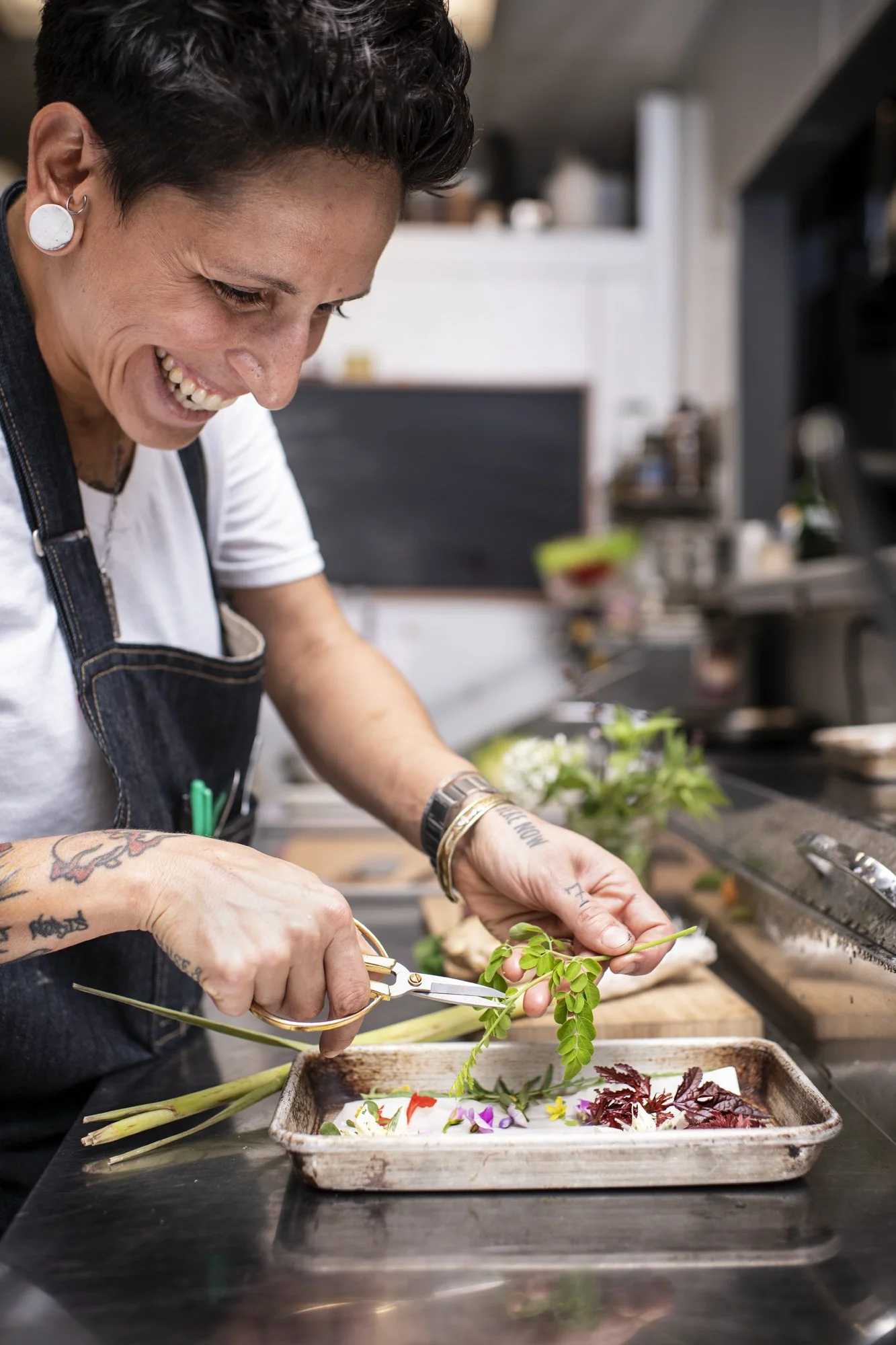 A chef with tattoos and earrings is preparing floral decorations in a kitchen, using scissors to trim greenery over a tray with colorful flowers and leaves.