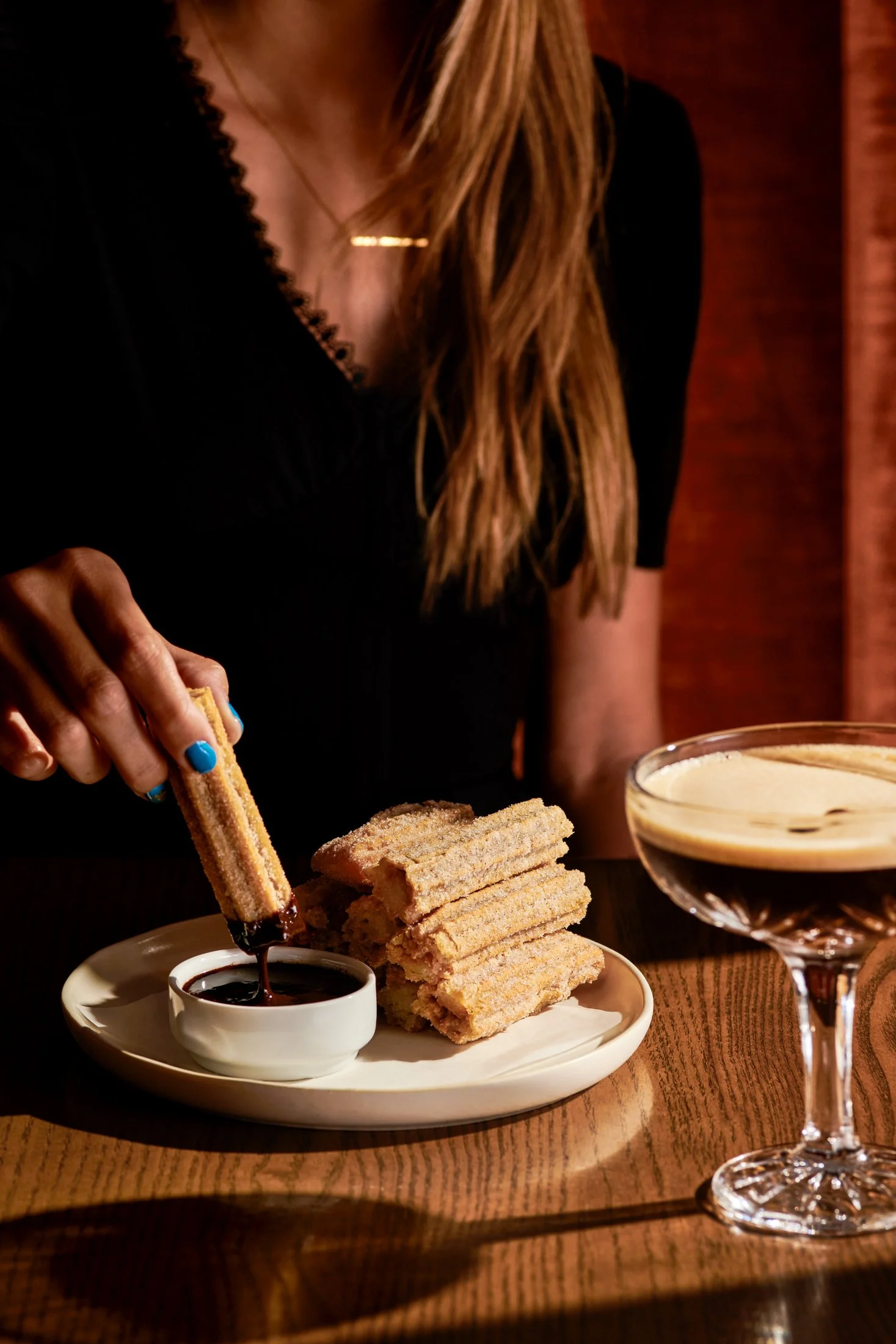 Person dipping a churro into a cup of chocolate sauce on a plate with stacked churros and a cocktail glass with a creamy drink on a wooden table.
