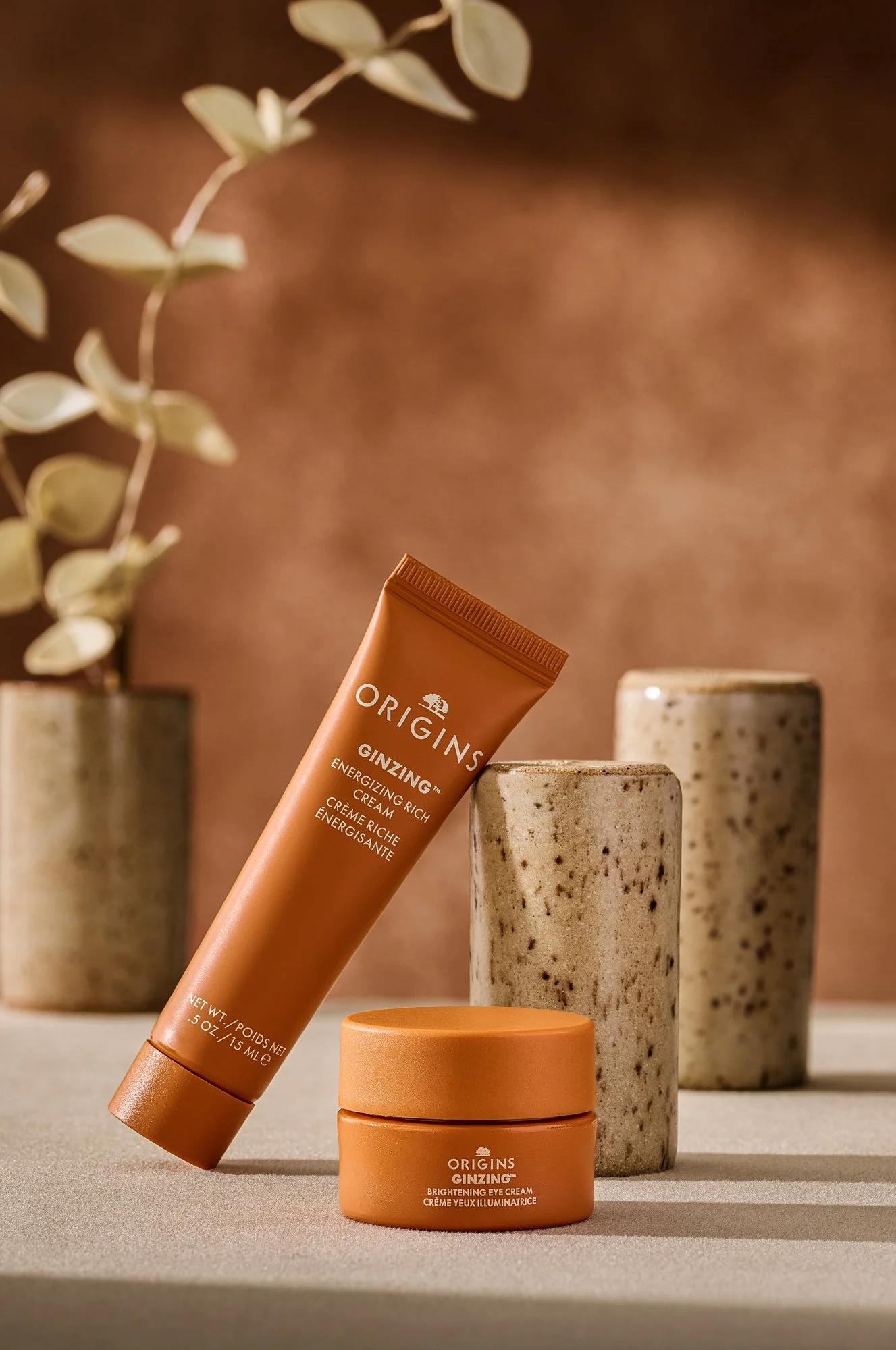 A warm scene of Origins beauty products on a soft surface with ceramic bud vase in background, copper wall behind with shadows. Product beauty photography by Nutmeg Imageworks.