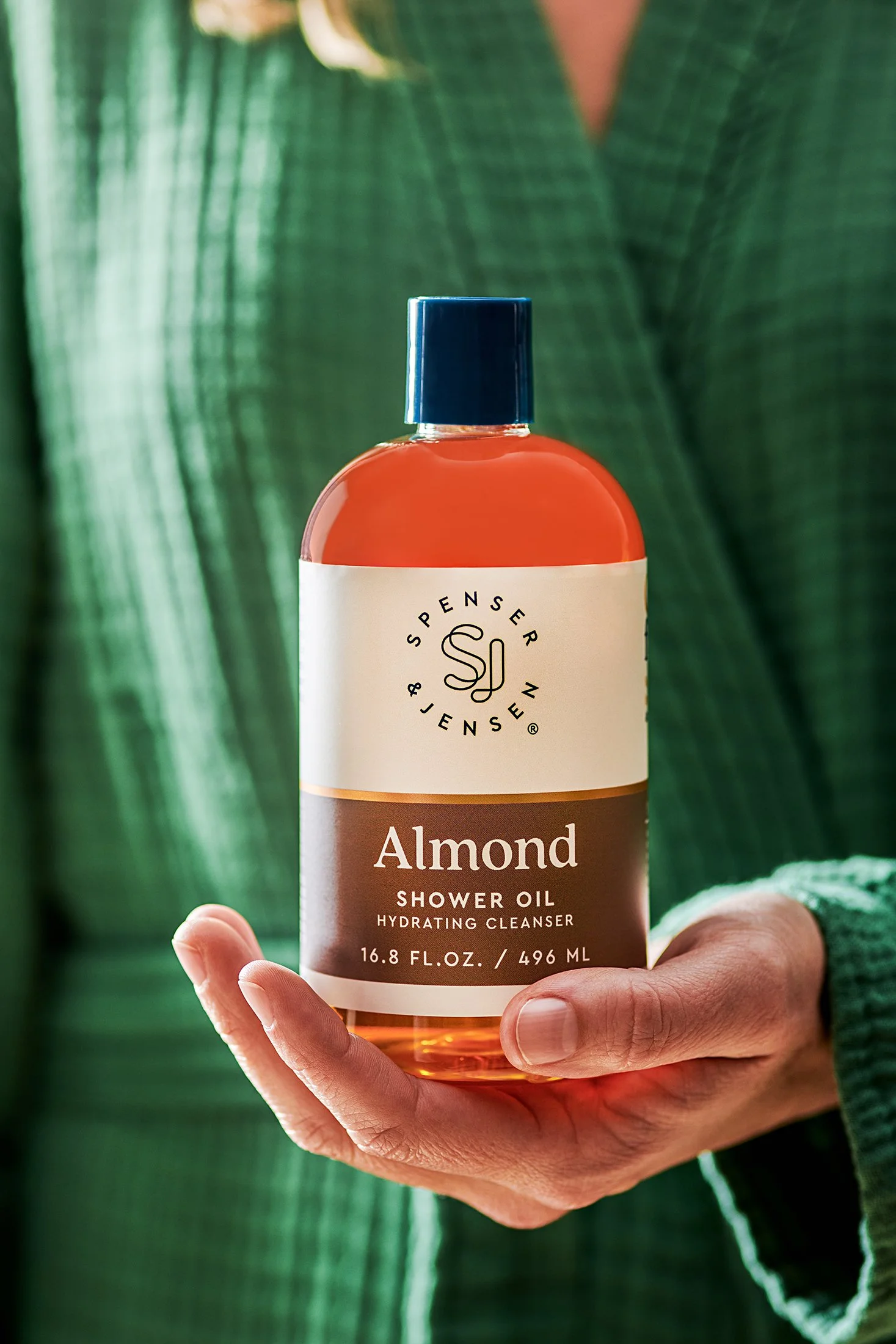 A hand holding a bottle of almond shower oil with a white label and blue cap, with green fabric in the background.